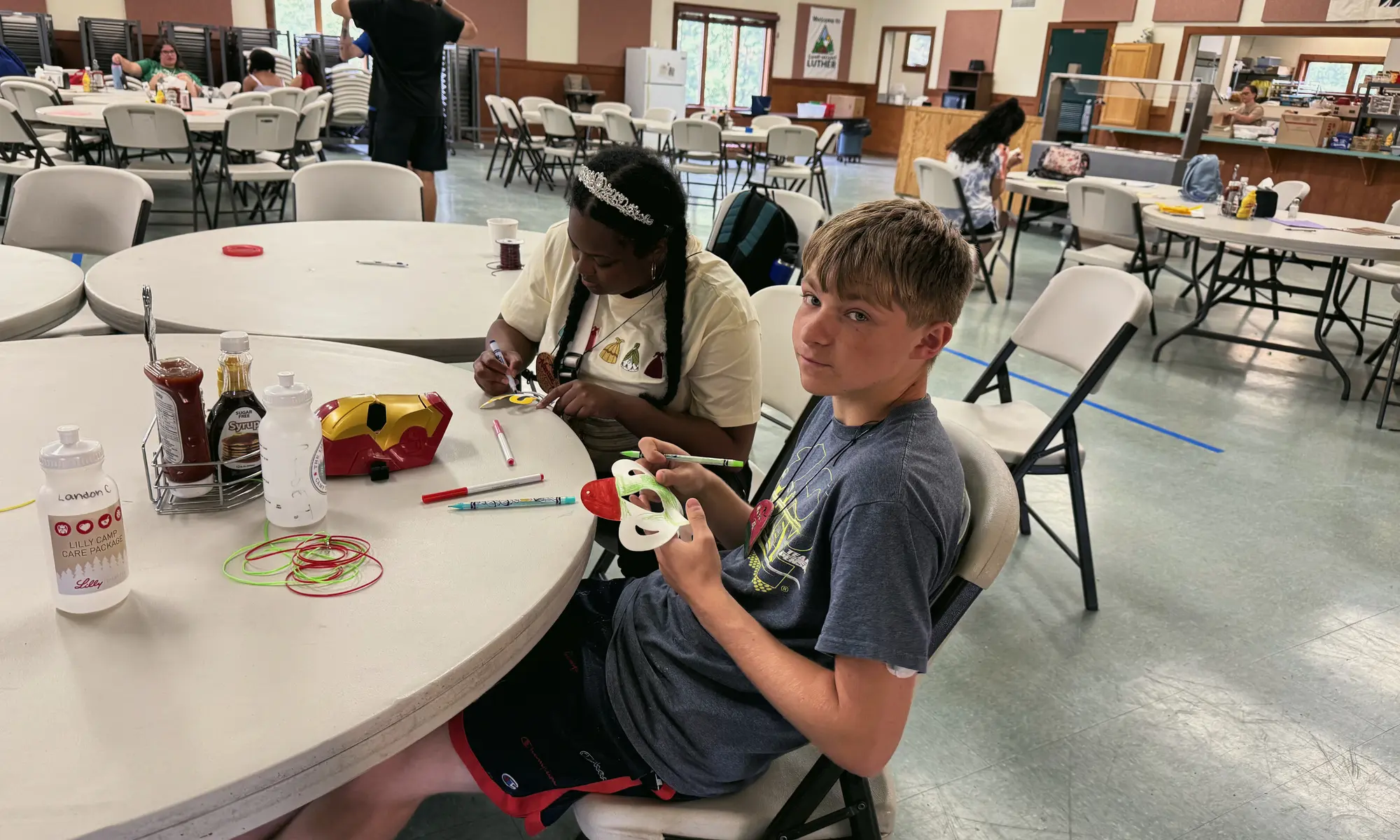 A boy shows his craft project at a round table in a busy hall, with a girl working beside him