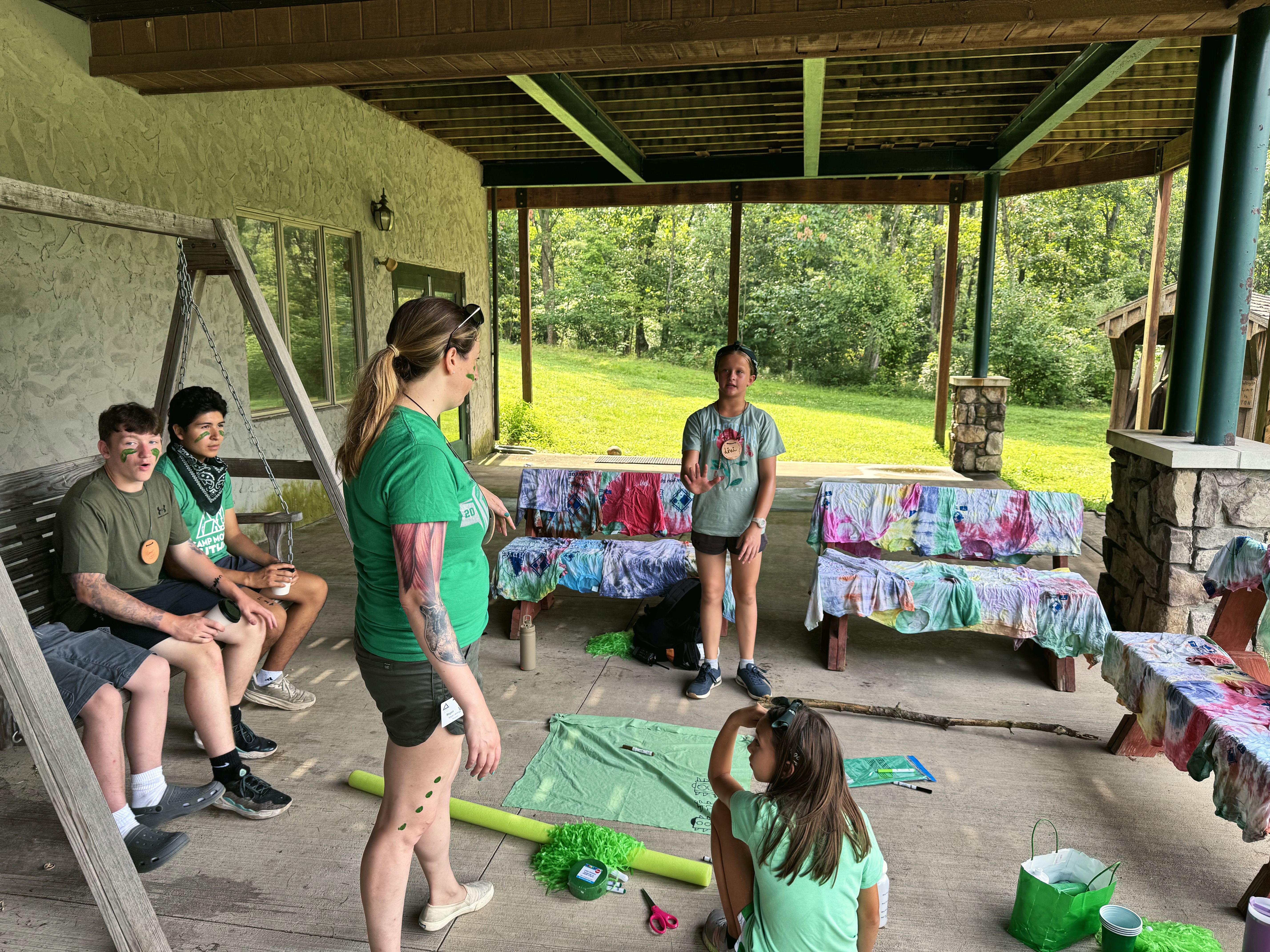 A counselor with a group of campers on a covered porch, with tie-dye shirts drying on the benches.