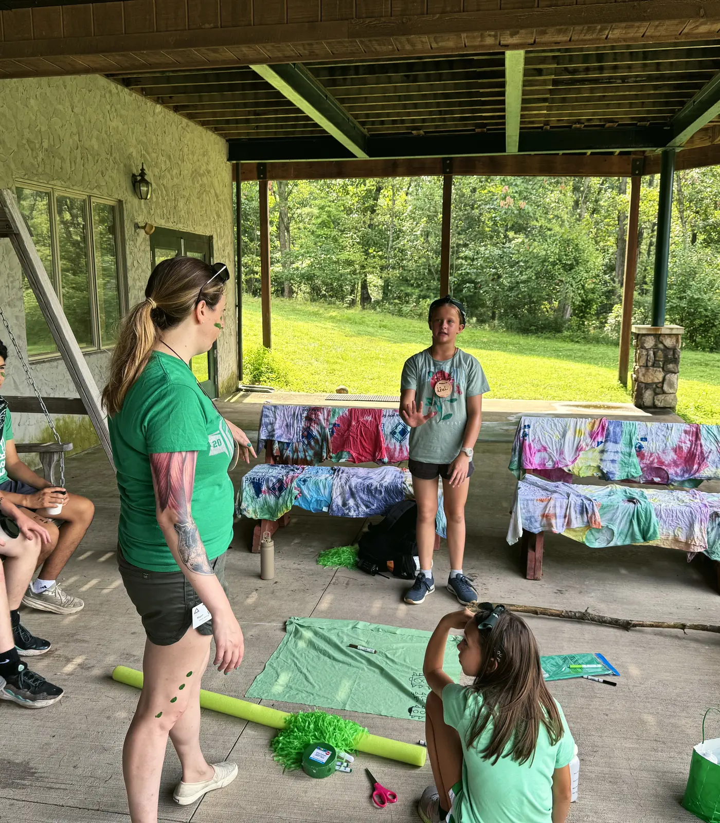 A counselor with a group of campers on a covered porch, with tie-dye shirts drying on the benches.