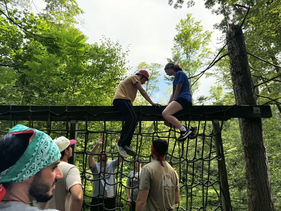 Campers climb over a log beam on the ropes course in the woods.