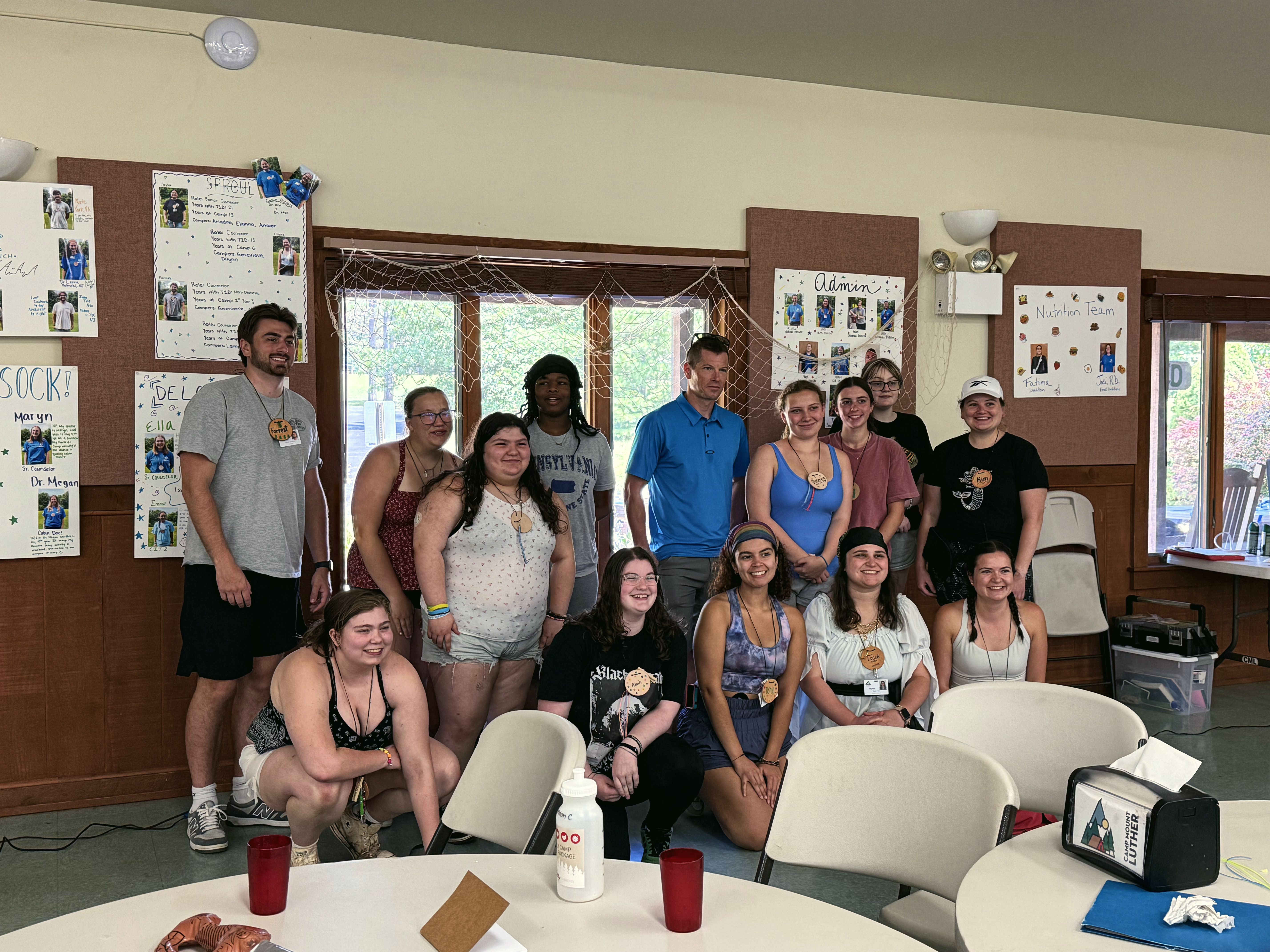 A group of young adults smile together for a photo in a camp hall.