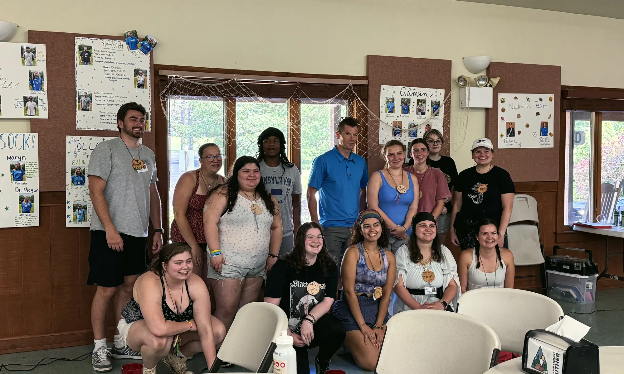 A group of young adults smile together for a photo in a camp hall.