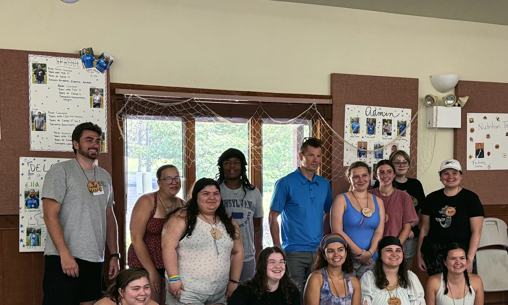 Olympic skier and fellow T1Der Kris Freeman, wearing a blue polo shirt, poses in the center of a group of young adult camp staff with wooden name tags as they smile together in the camp hall.