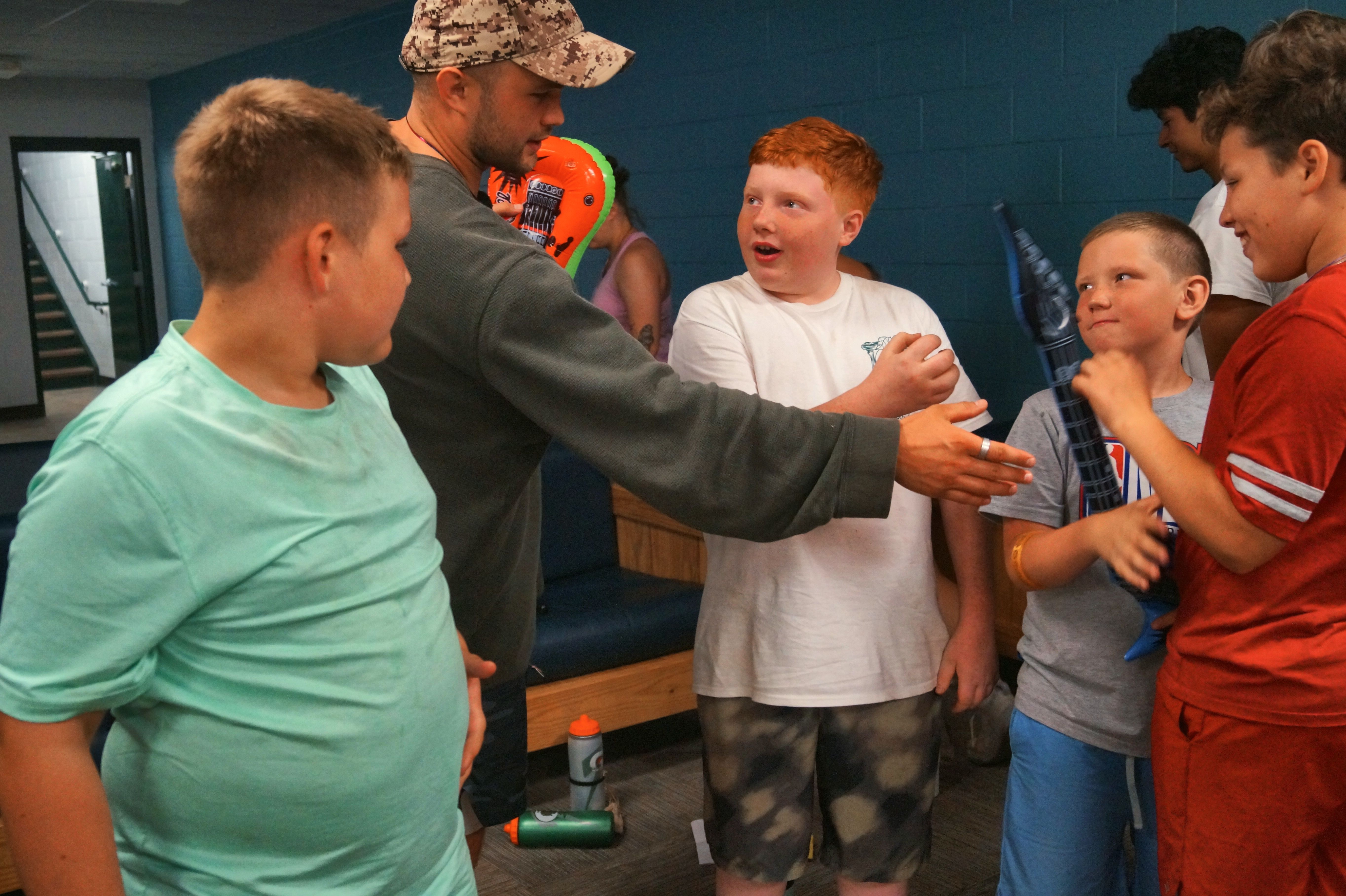 A counselor in a camouflage hat talks with a group of excited boys indoors at camp.