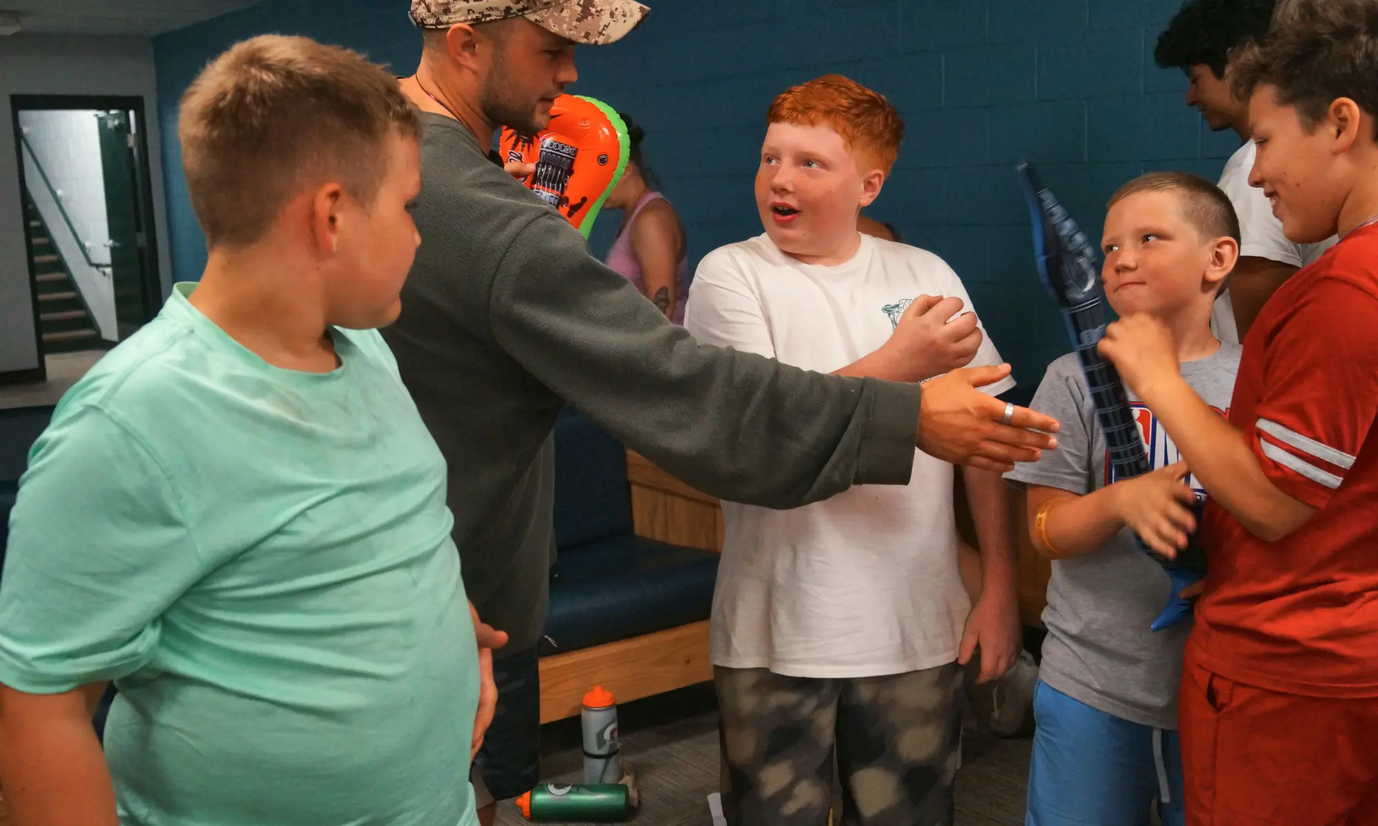 A counselor in a camouflage hat talks with a group of excited boys indoors at camp.
