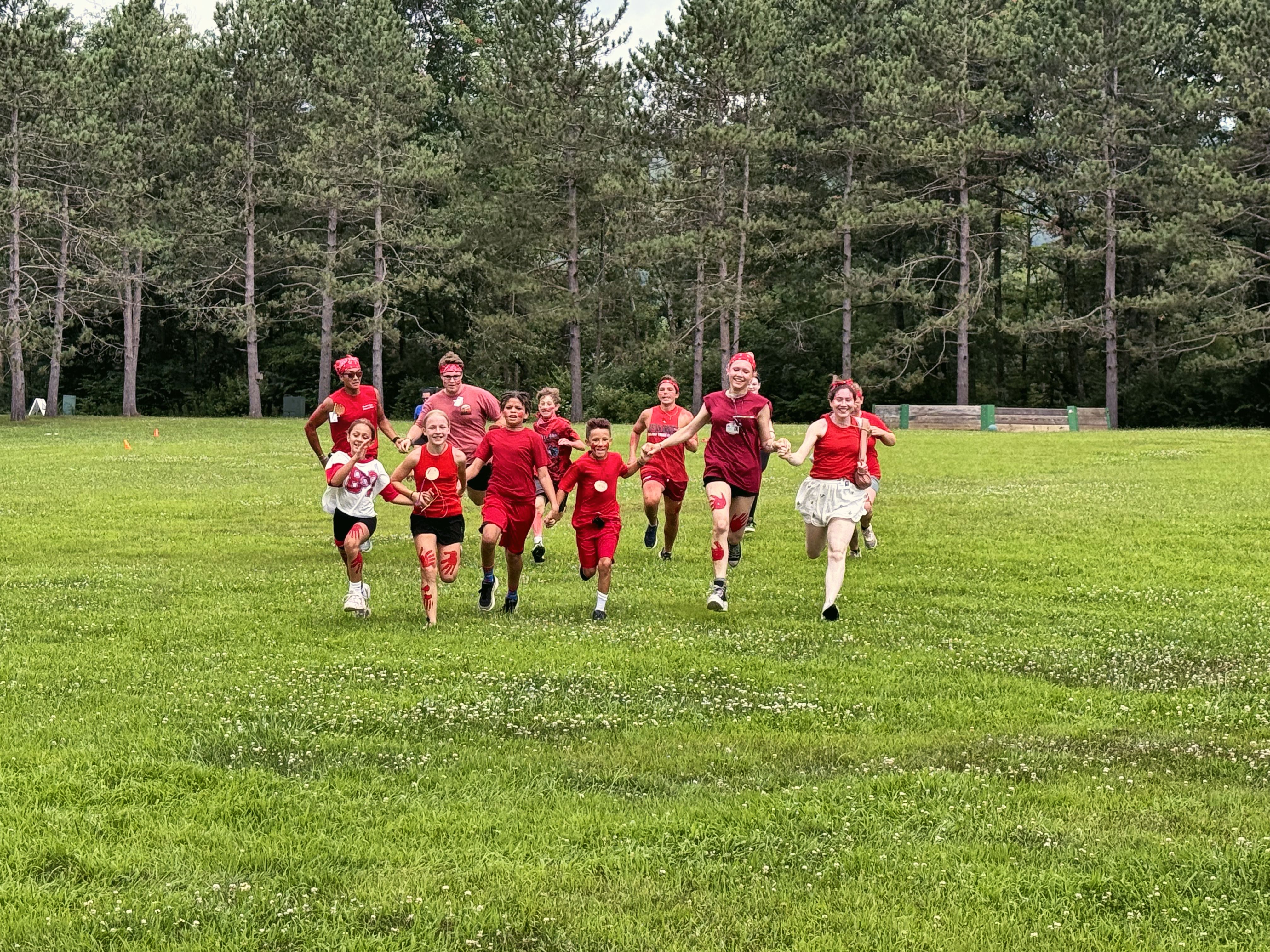 Young campers in red shirts run across a grassy field toward the camera, with tall pines behind them