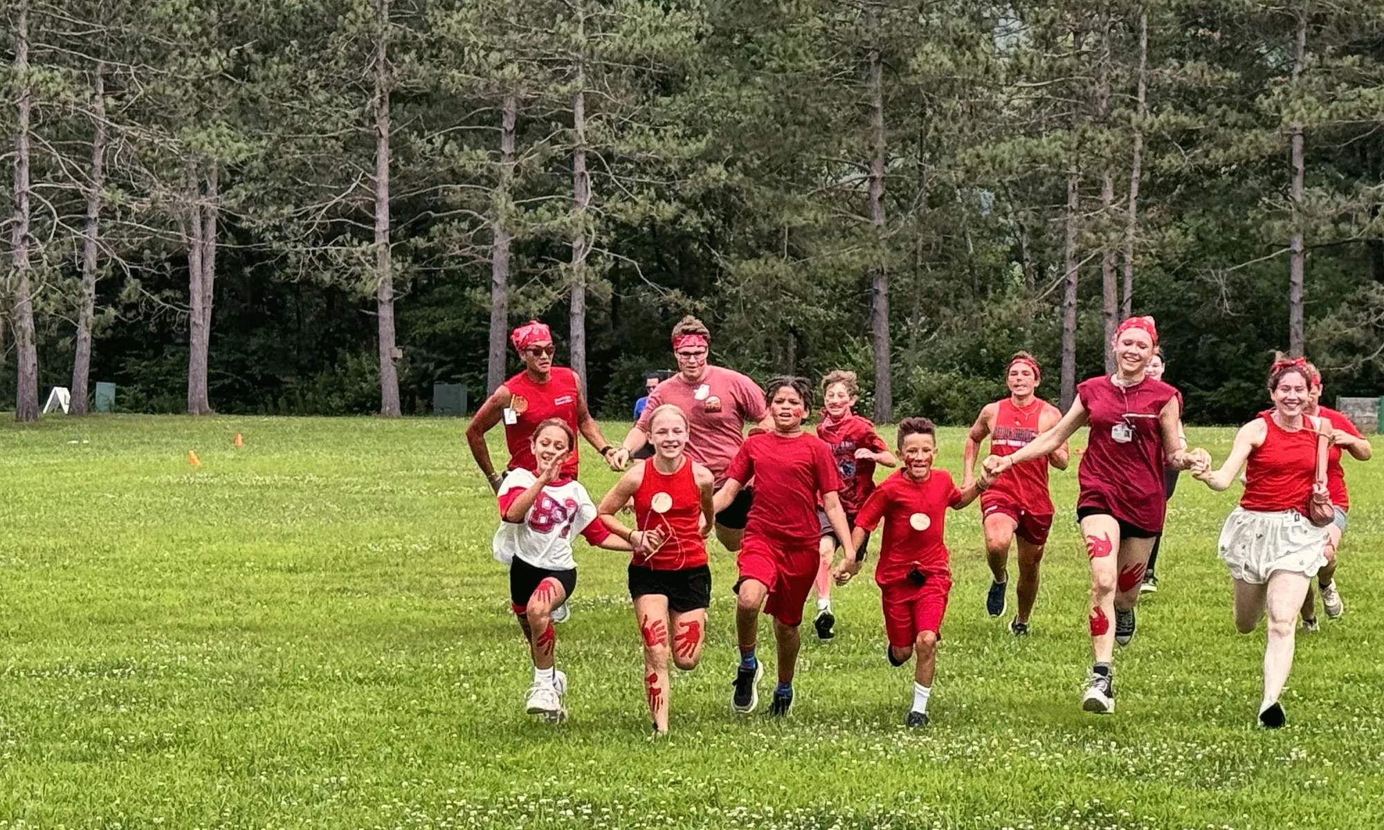 Young campers in red shirts run across a grassy field toward the camera, with tall pines behind them