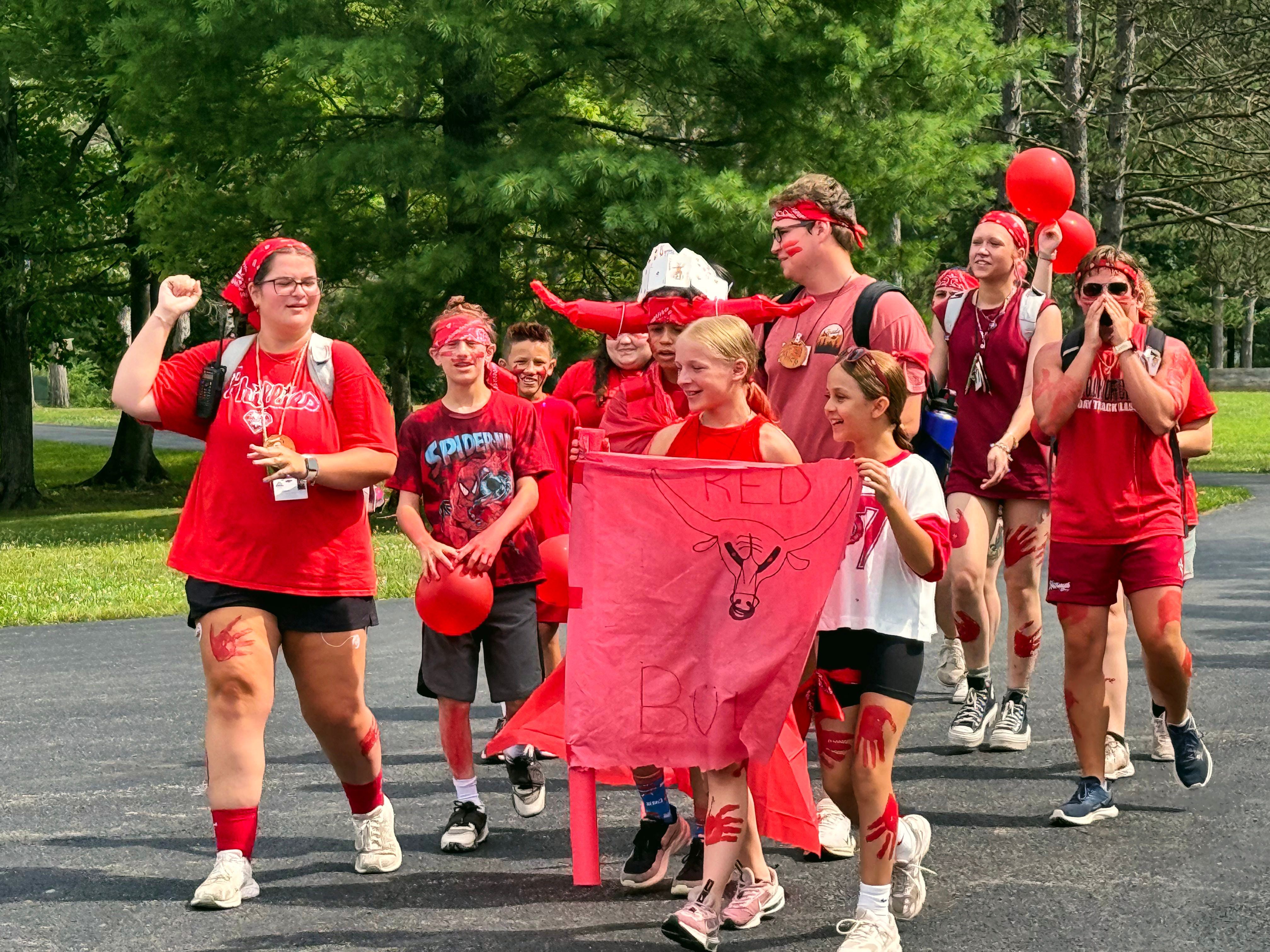 Campers in red cheer and march down a camp path with their team banner