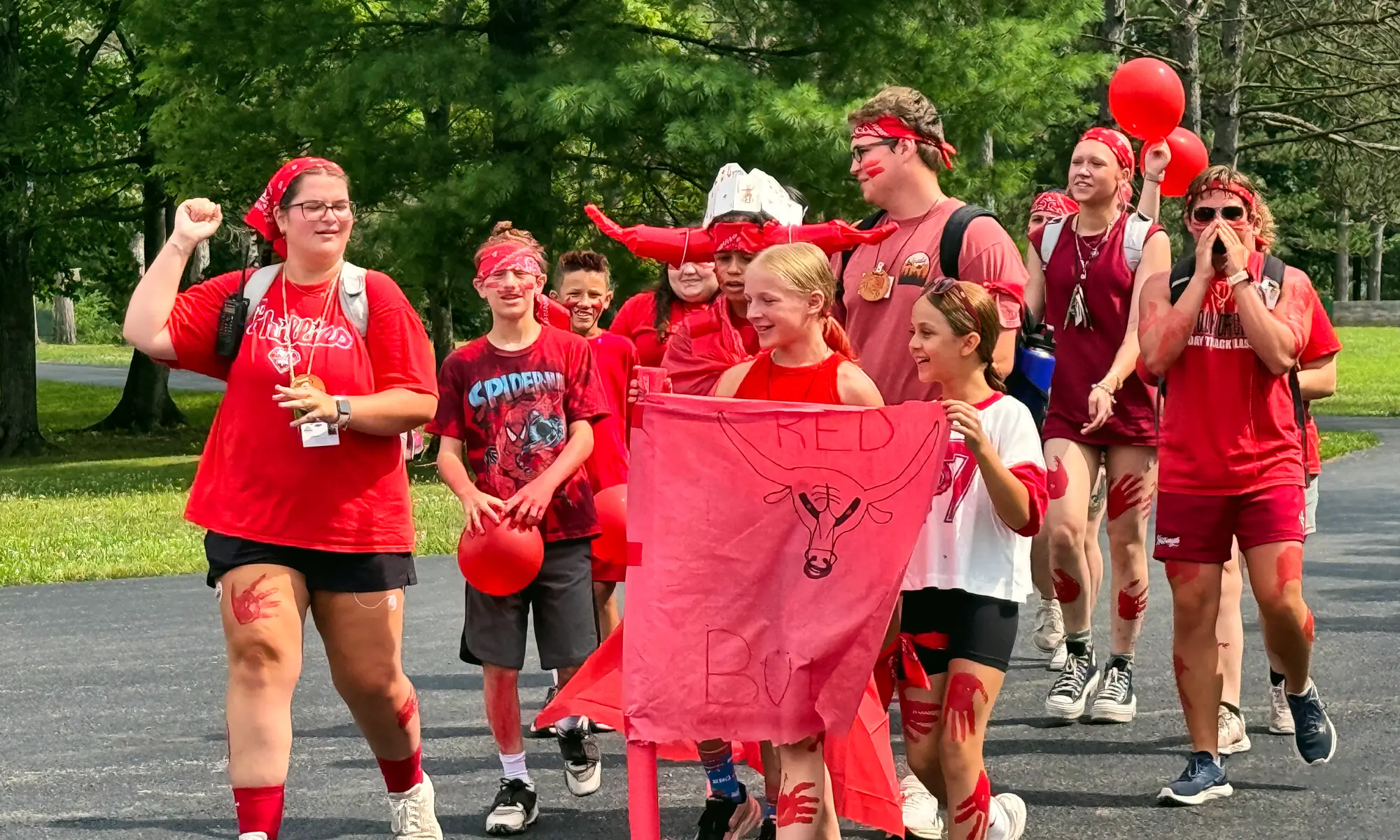Campers in red cheer and march down a camp path with their team banner