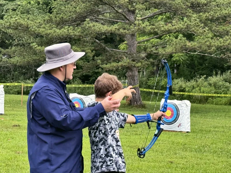 An adult shows a young camper how to hold a bow at the archery range