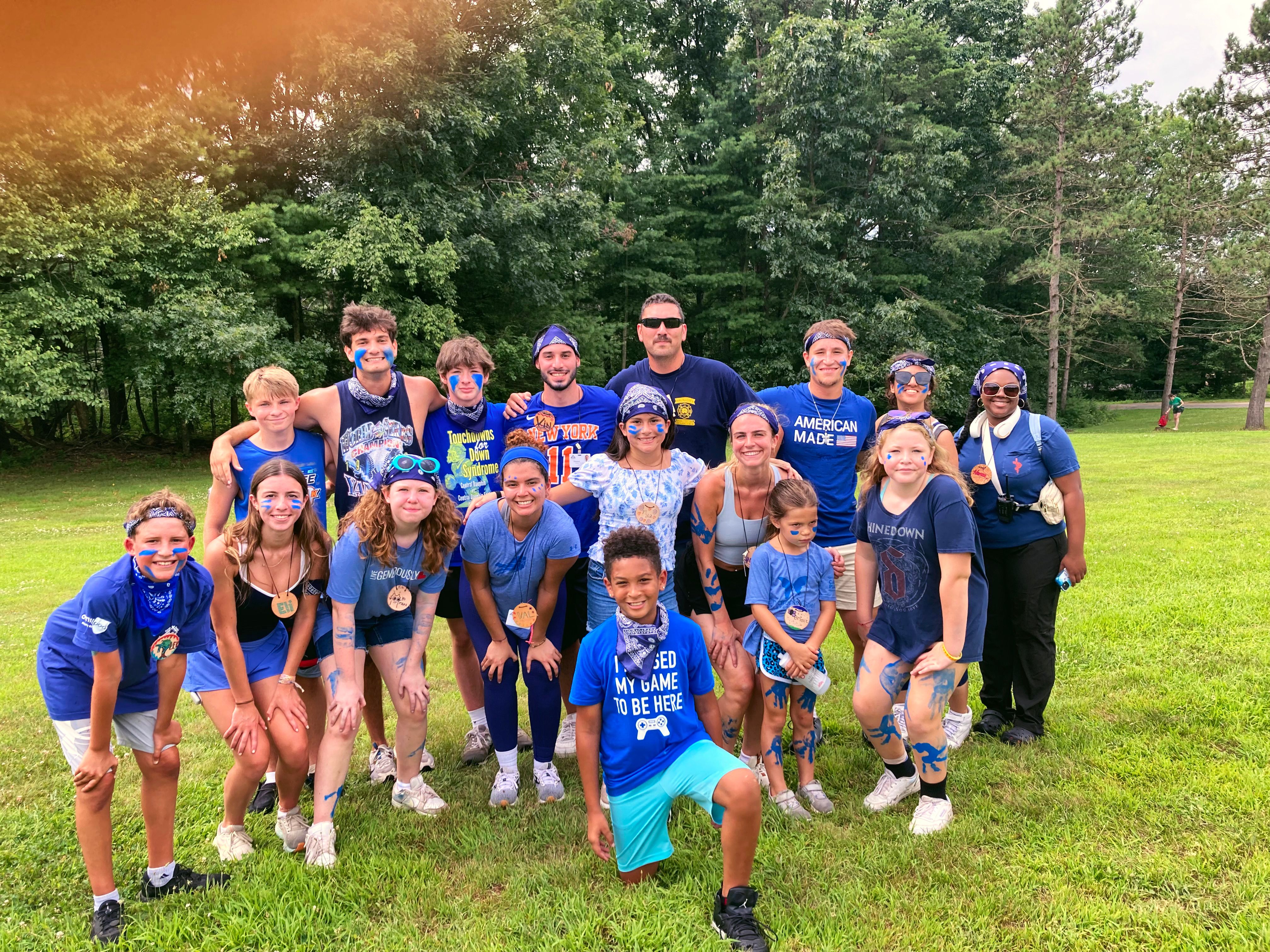 Teens and adults in matching blue shirts, some with blue face paint, pose together on a grassy field at camp