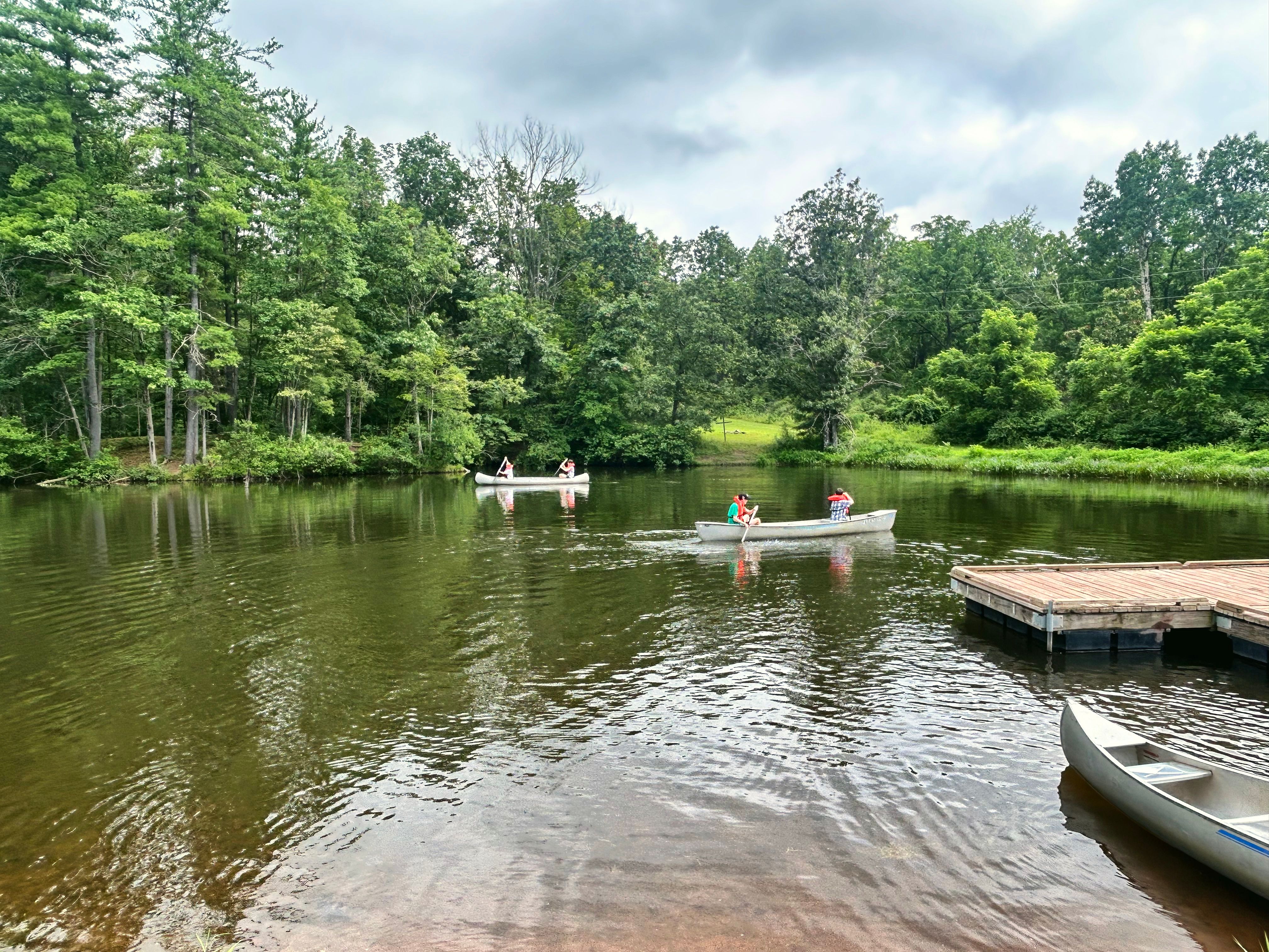 Canoes glide across a calm lake with a wooden dock, surrounded by trees.