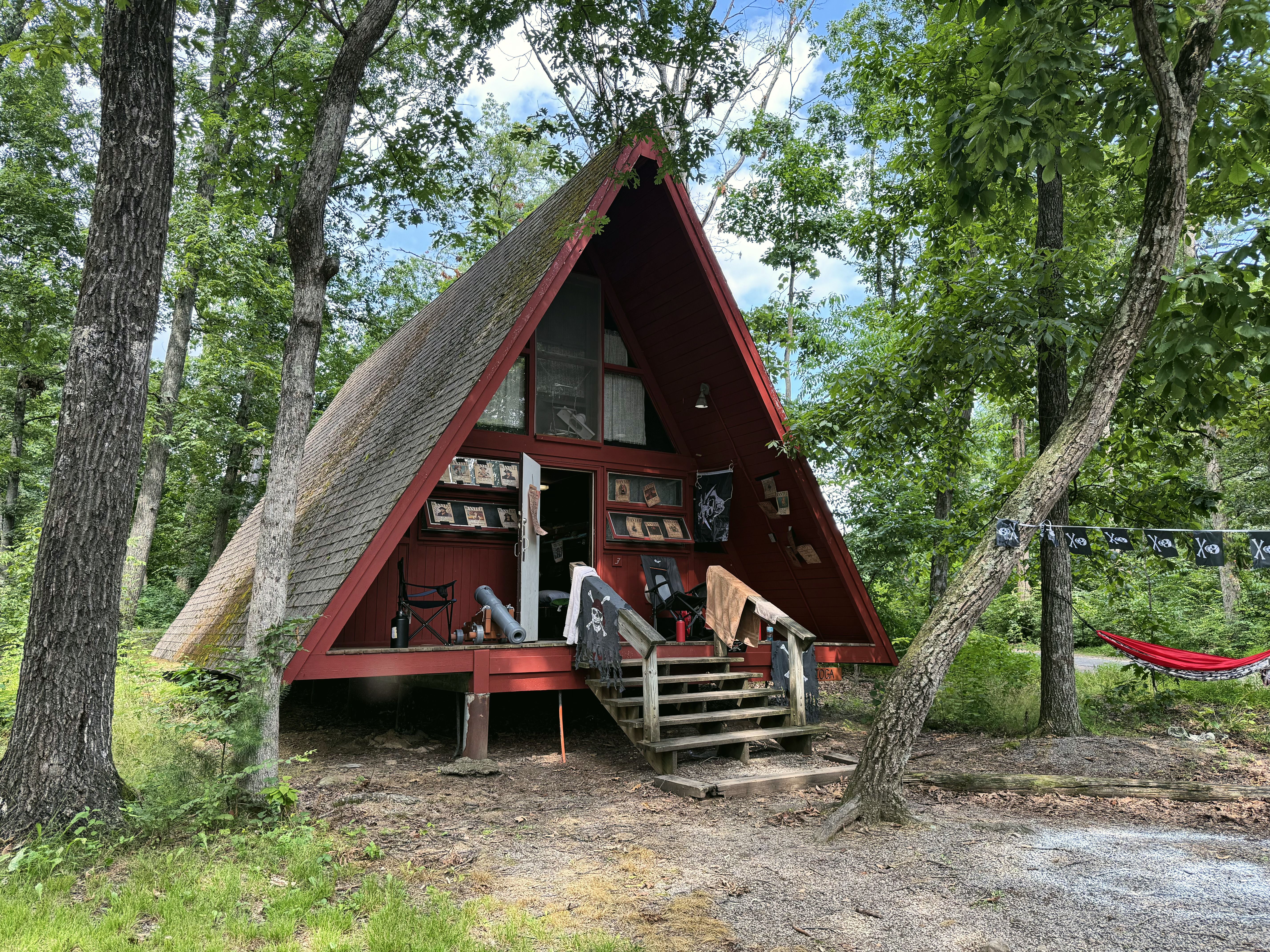 A red A-frame cabin with a wooden porch among tall trees, with a hammock beside it