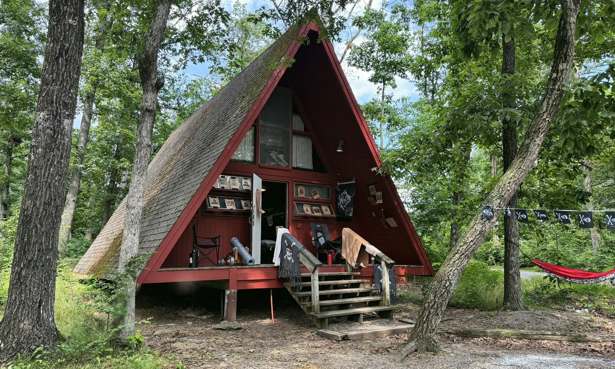 A red A-frame cabin with a wooden porch among tall trees, with a hammock beside it