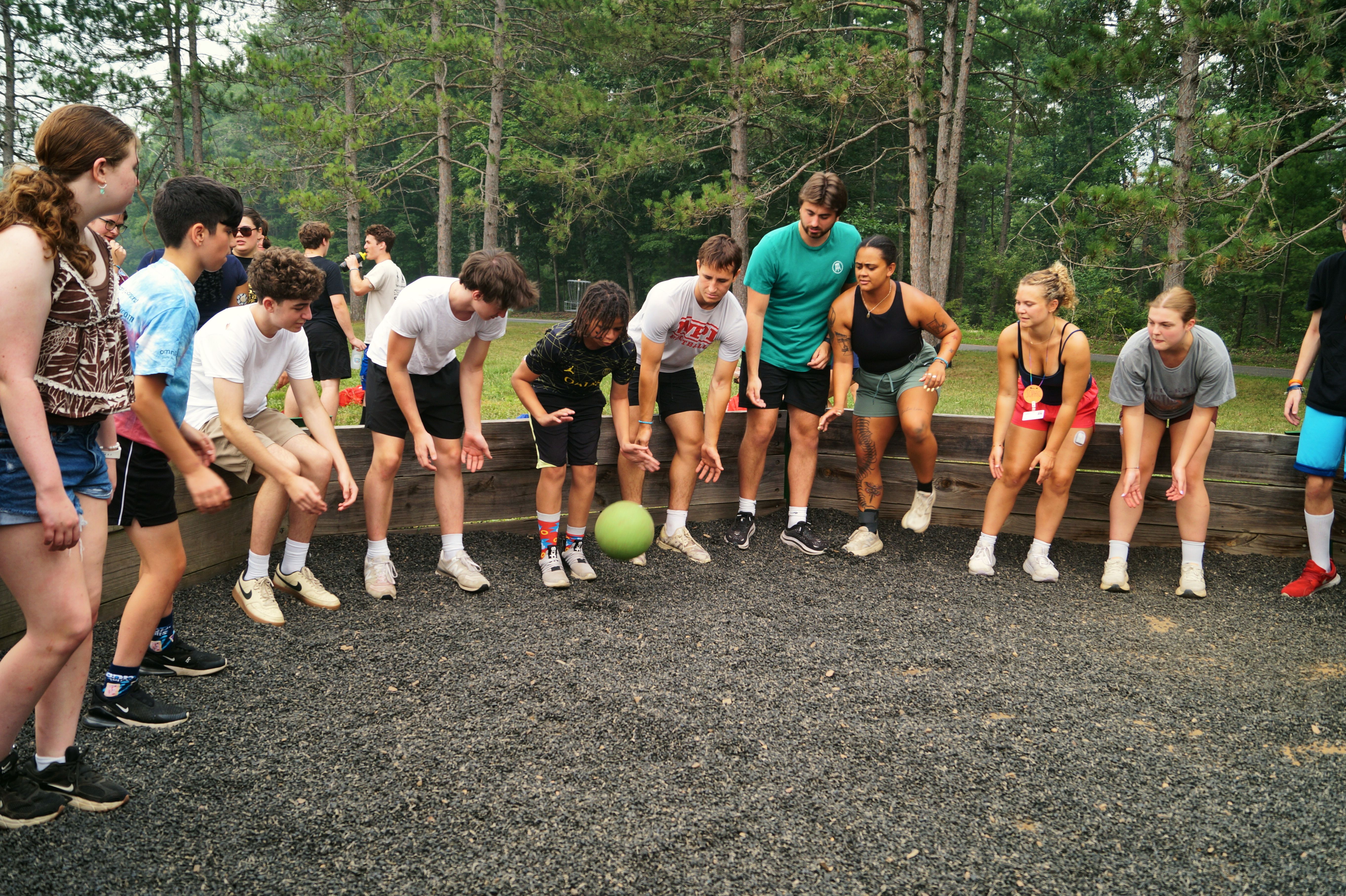 Young people stand around a wooden ball pit and play a game with a green ball.
