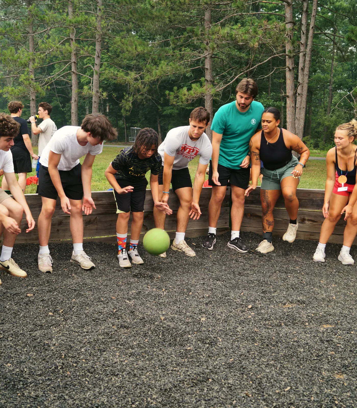 Young people stand around a wooden ball pit and play a game with a green ball.