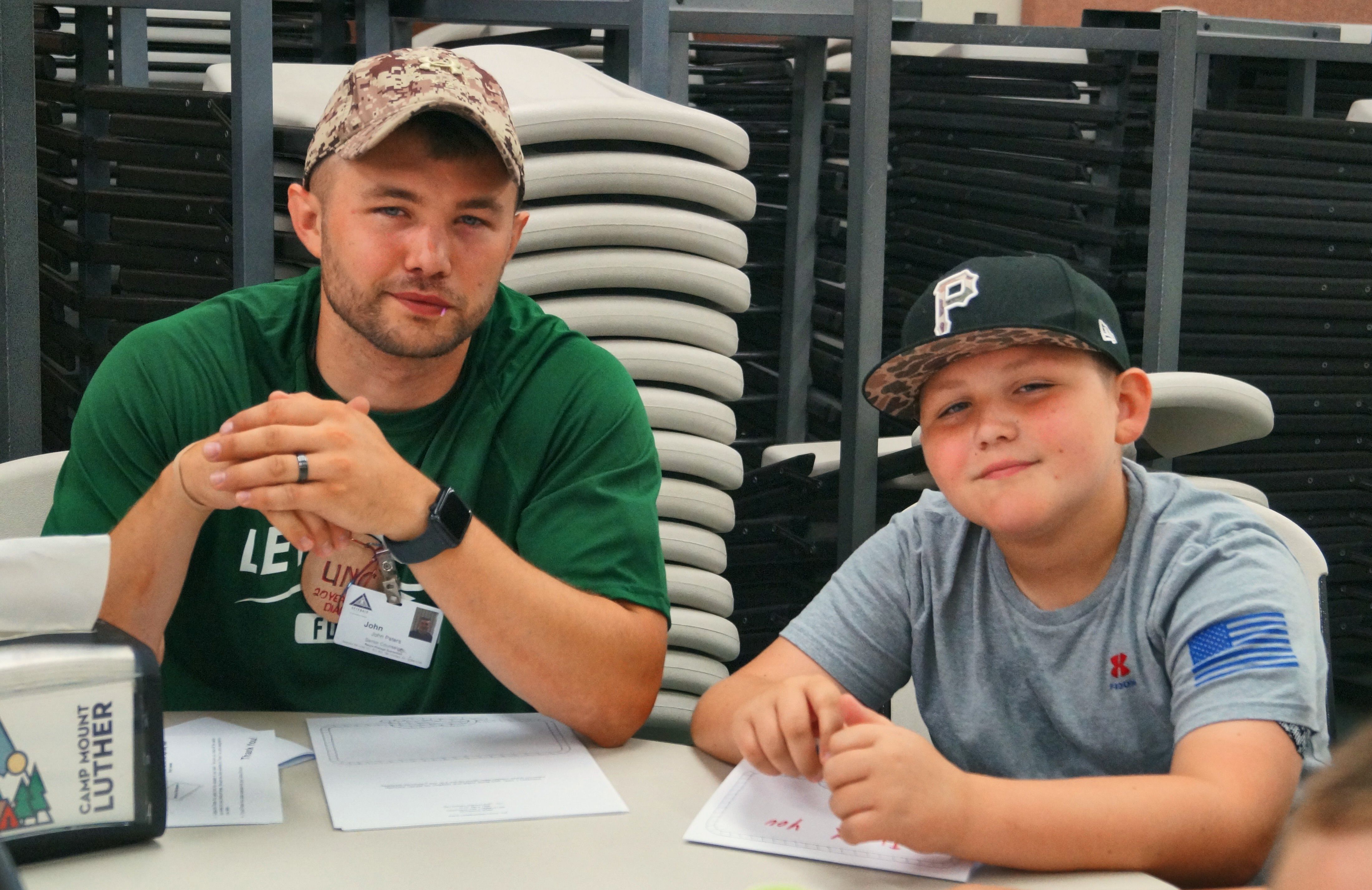 A male counselor in a green shirt and a camper in a baseball cap sit together at a table.