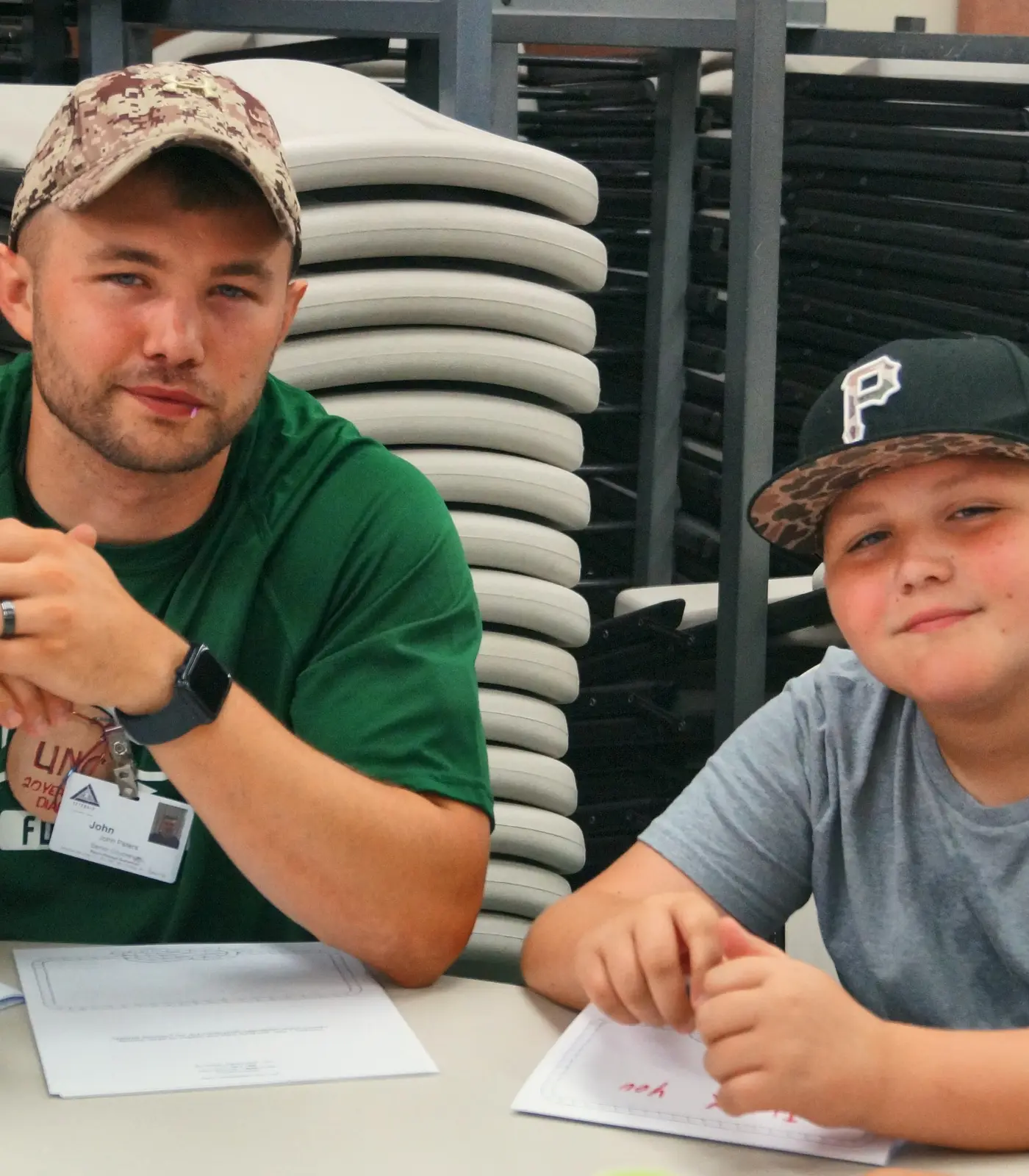 A male counselor in a green shirt and a camper in a baseball cap sit together at a table.