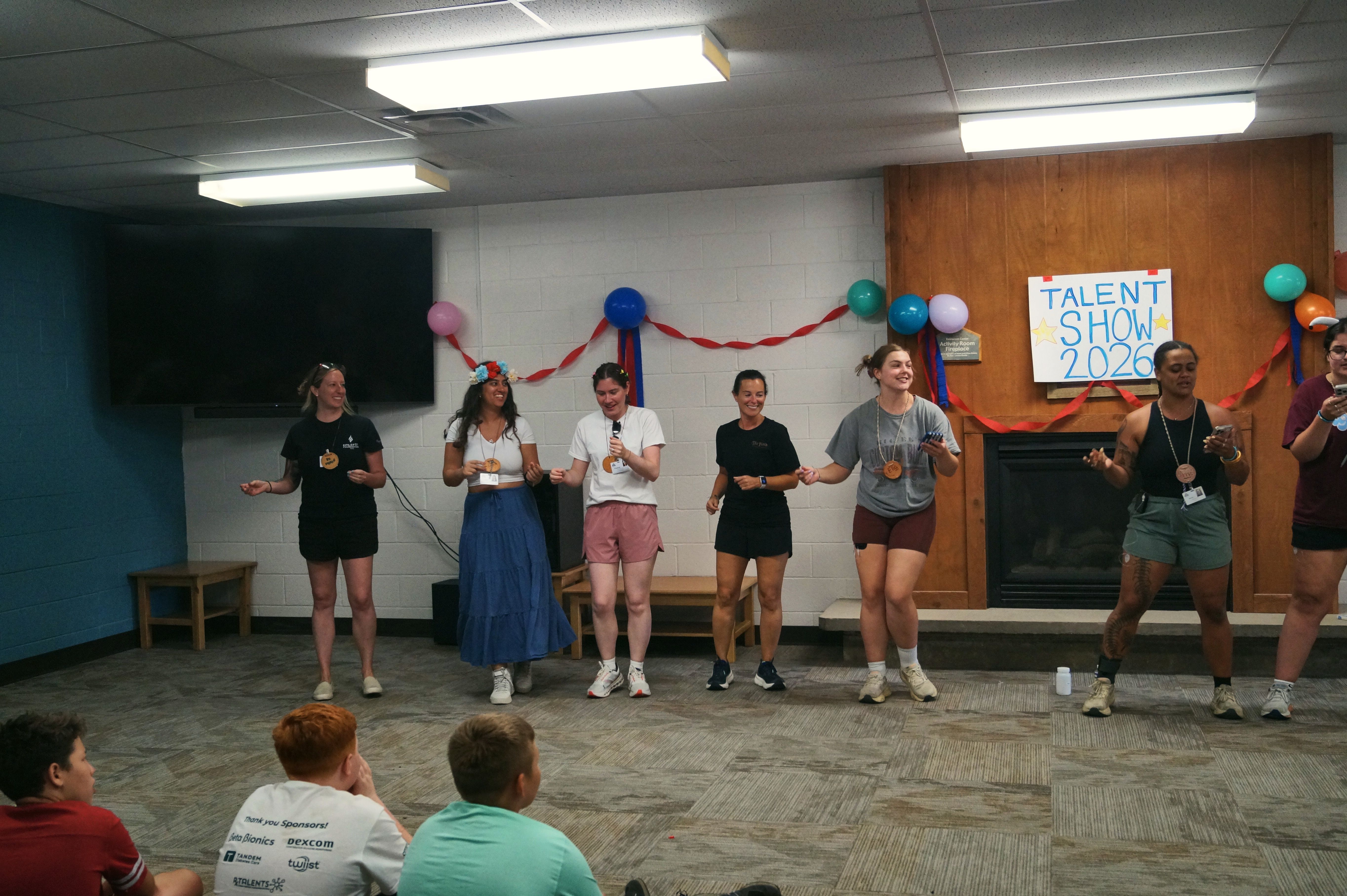 A group of young women perform on a stage at a camp talent show while the audience watches