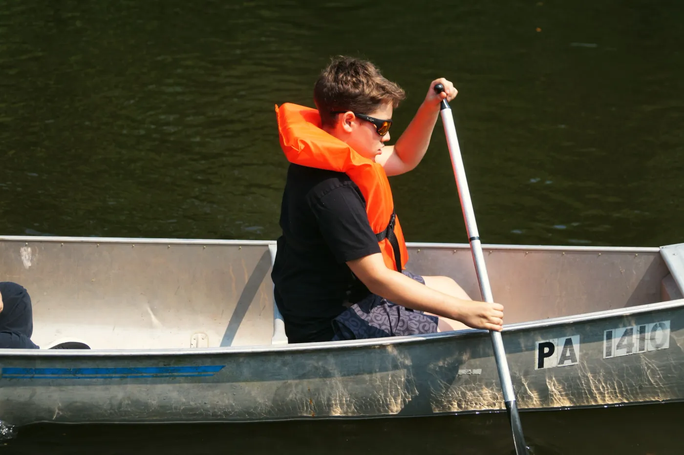 A young camper in an orange life vest and sunglasses paddles a canoe on calm water
