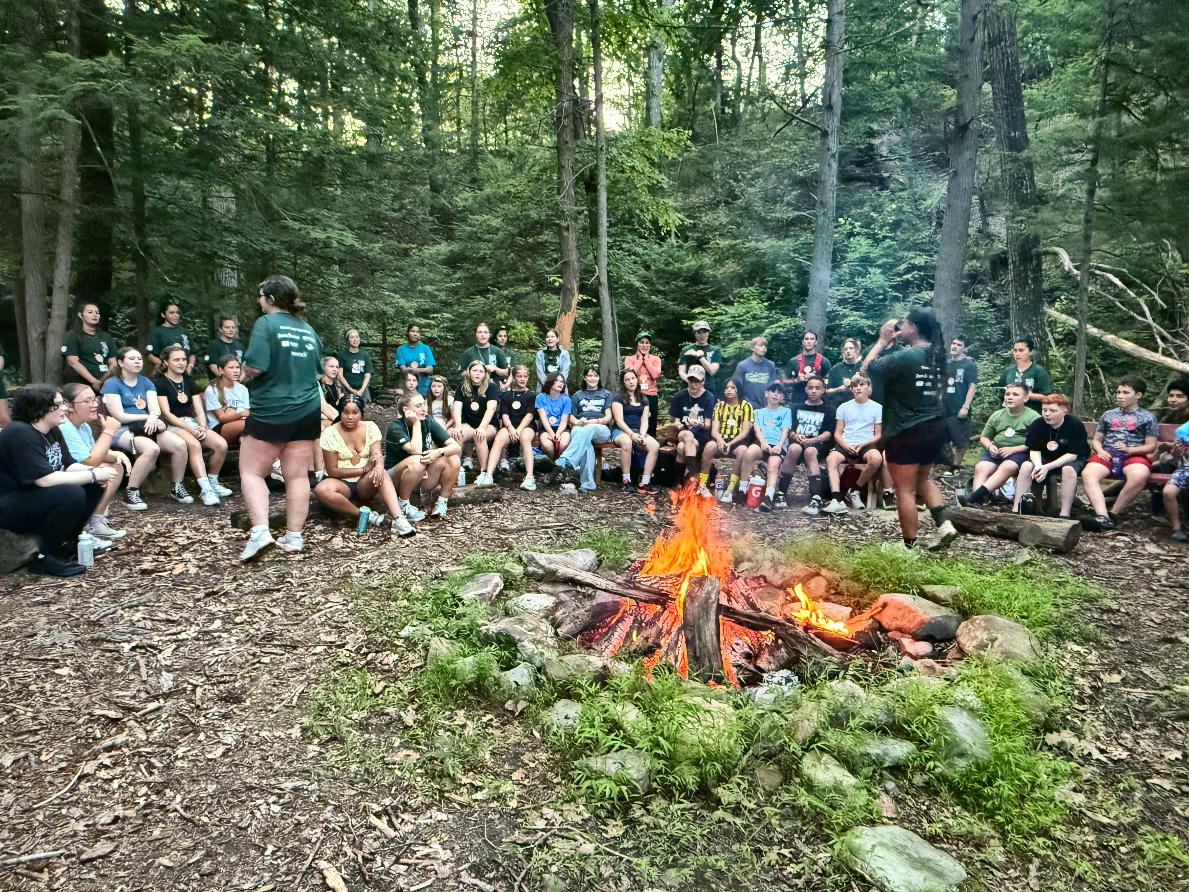 Teen campers sit on benches around a campfire in the woods while a counselor talks to the group