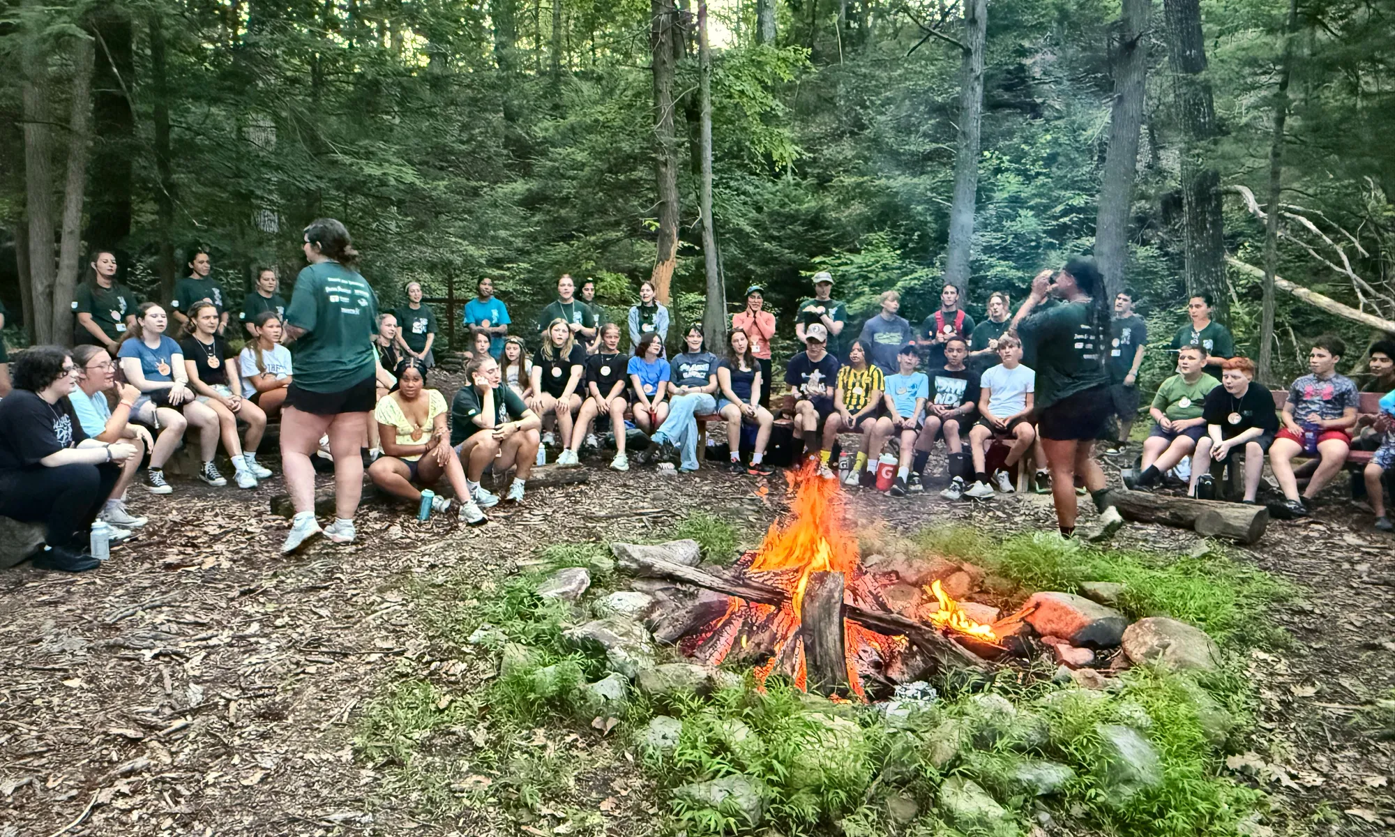 Teen campers sit on benches around a campfire in the woods while a counselor talks to the group