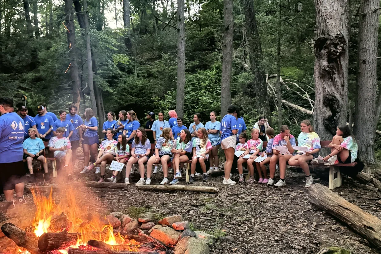 Campers and staff sit on logs and benches around a campfire in the woods