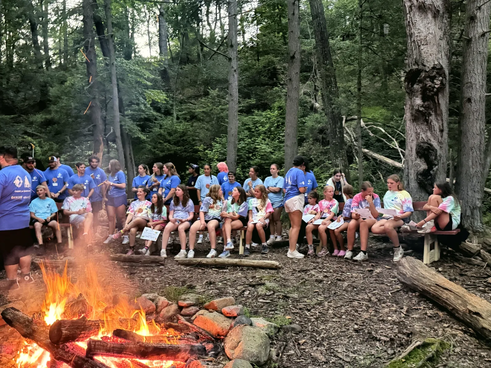Campers and staff sit on logs and benches around a campfire in the woods