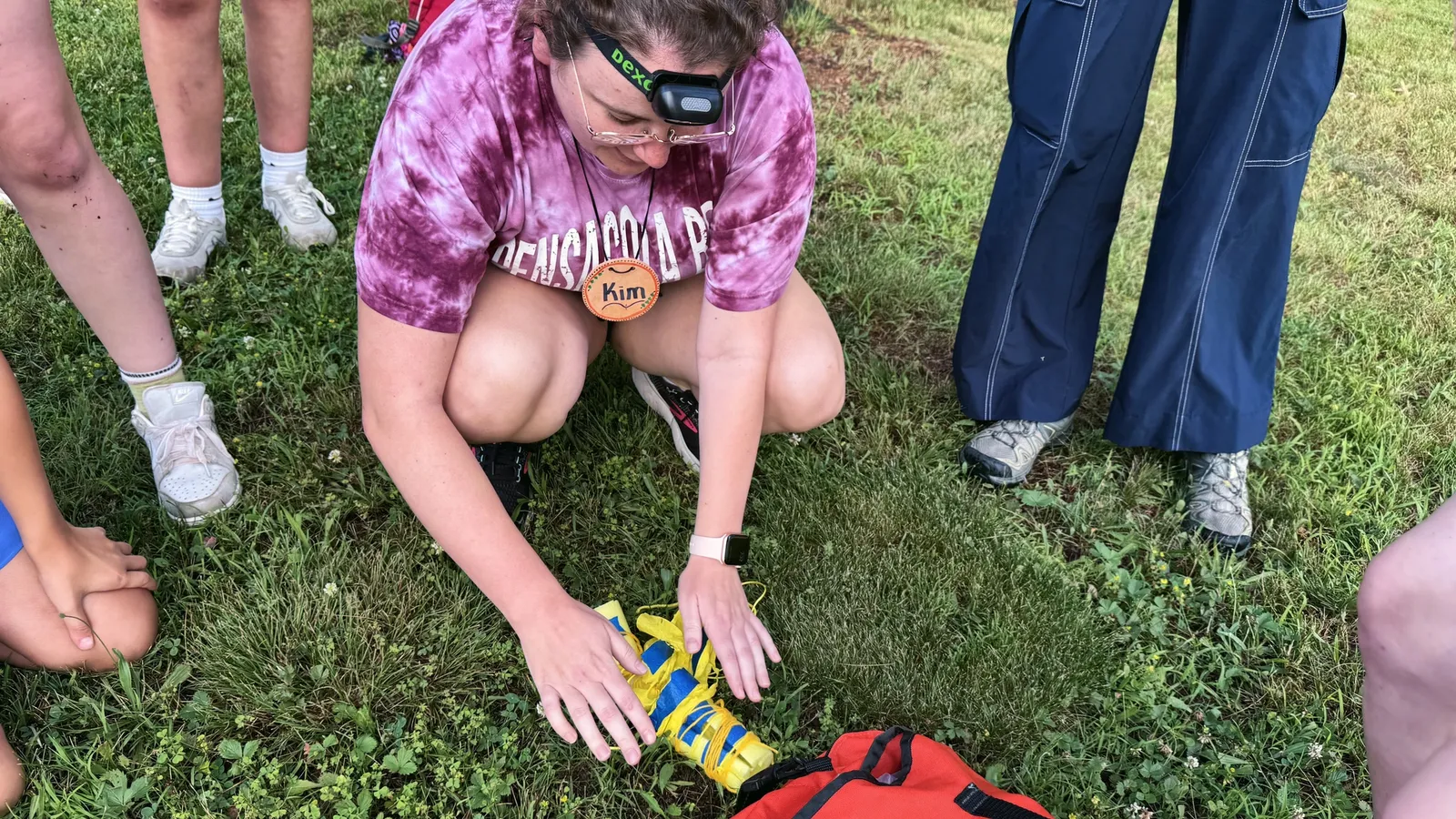 A staff member kneels on the grass and shows a first-aid splint from a red medical bag