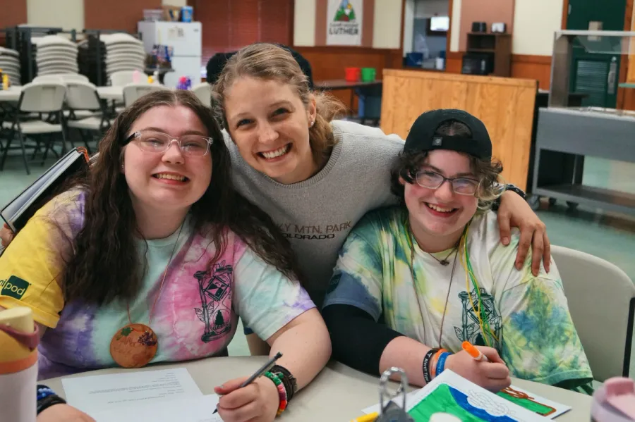 Three smiling campers sit together at a craft table. The camper in the middle has her arms around the other two.