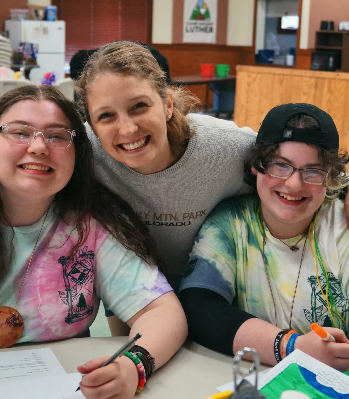 Three smiling campers sit close together at a craft table, one with her arms around the others