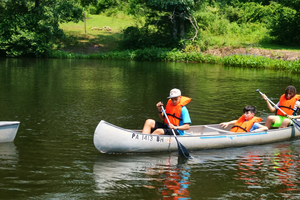 Three campers in orange life vests paddle a canoe on a calm lake