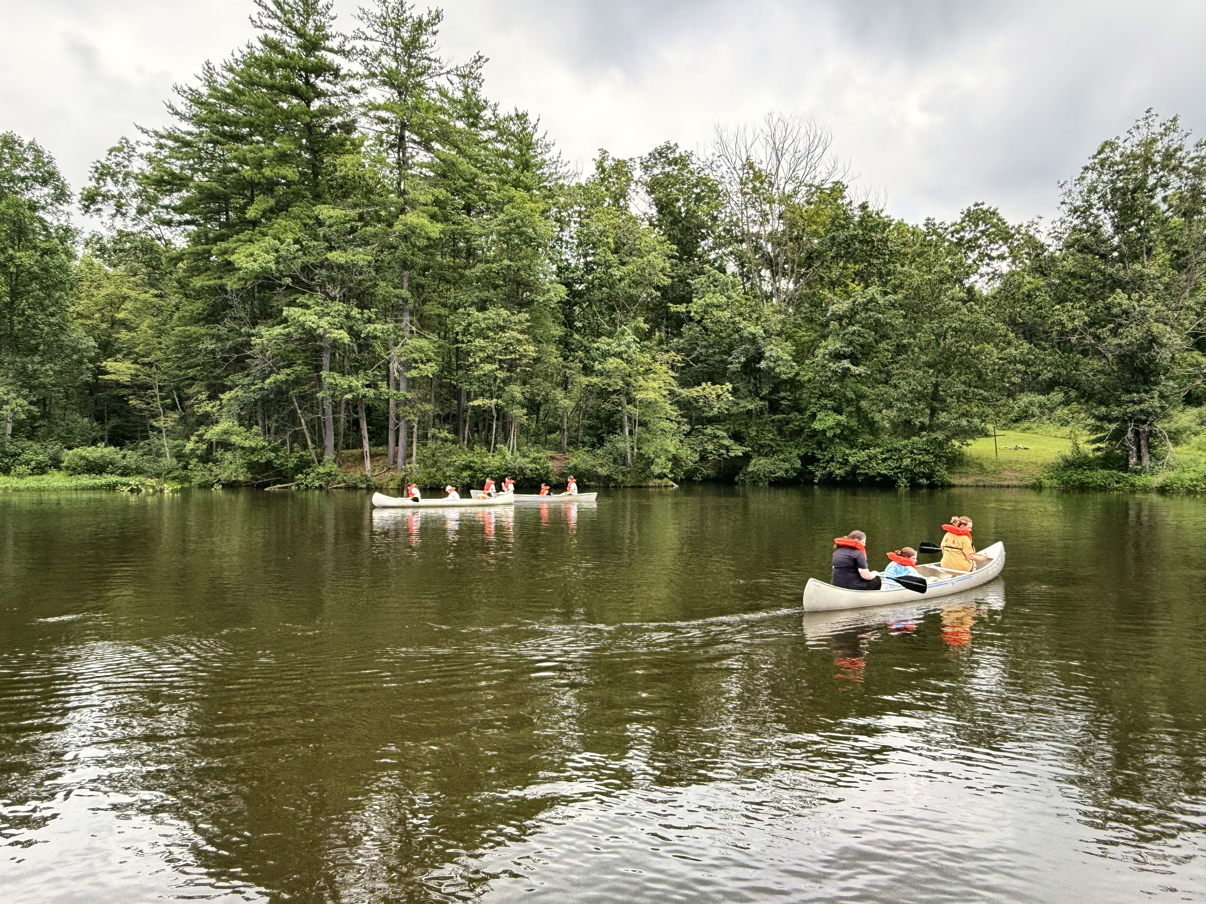 Campers in orange life vests paddle canoes across a calm, tree-lined lake at camp.