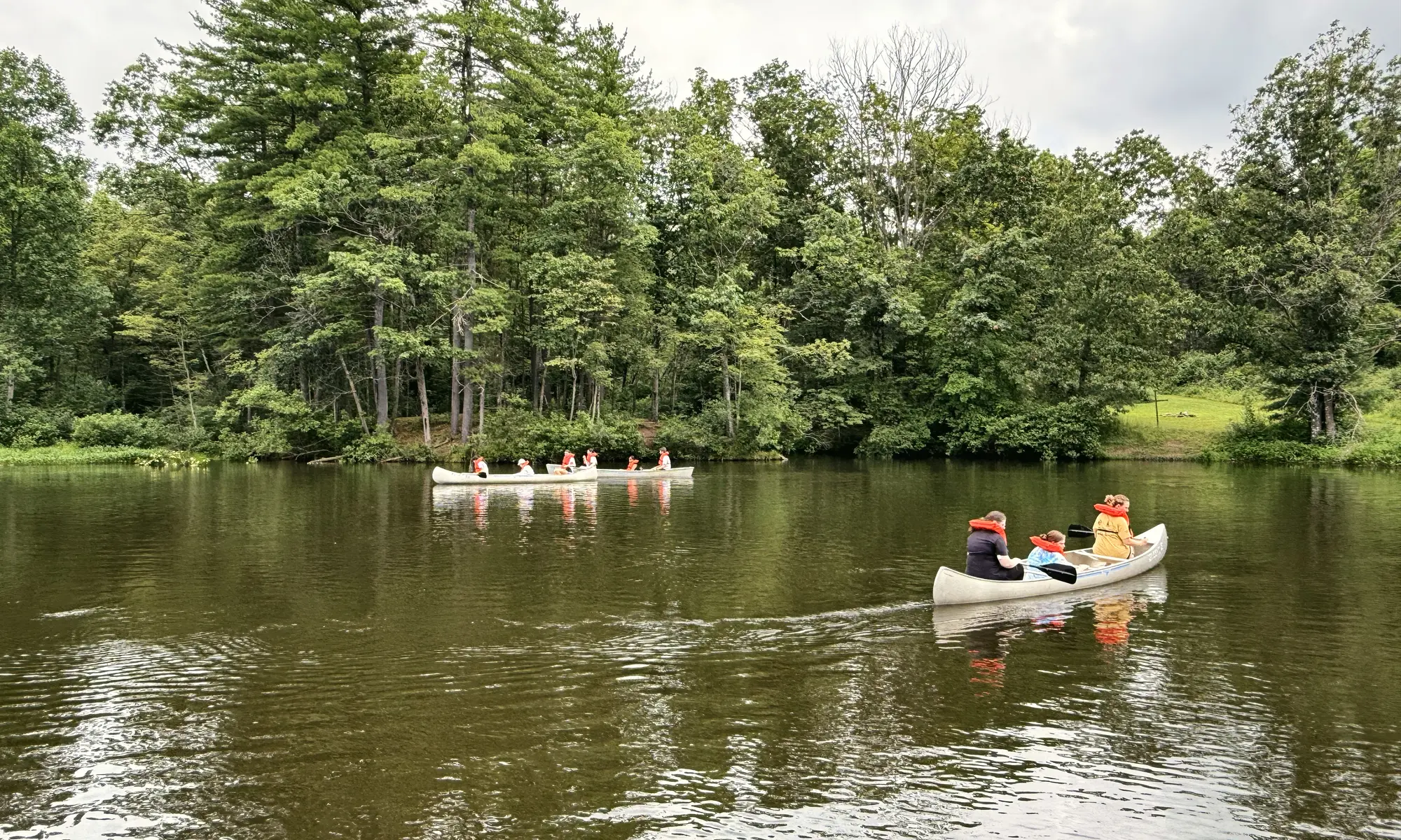Campers in orange life vests paddle canoes across a calm, tree-lined lake at camp.