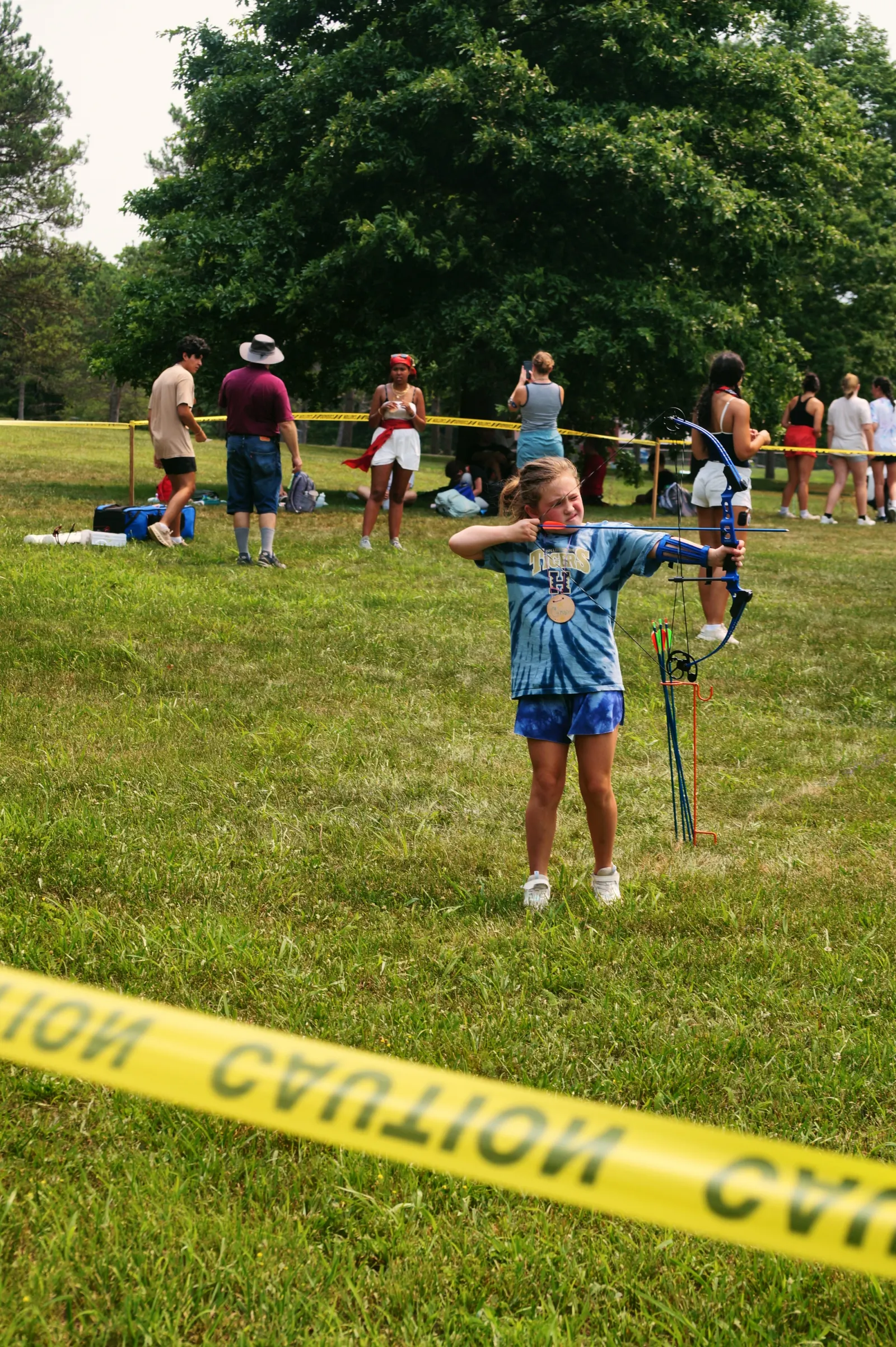 A girl in a tie-dye shirt aims a bow and arrow at a target on the archery field