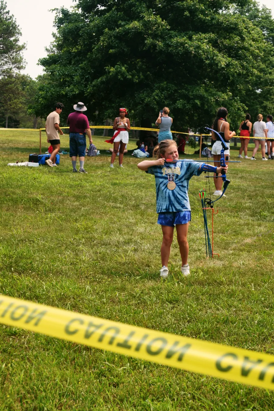A girl in a tie-dye shirt aims a bow and arrow at an archery target