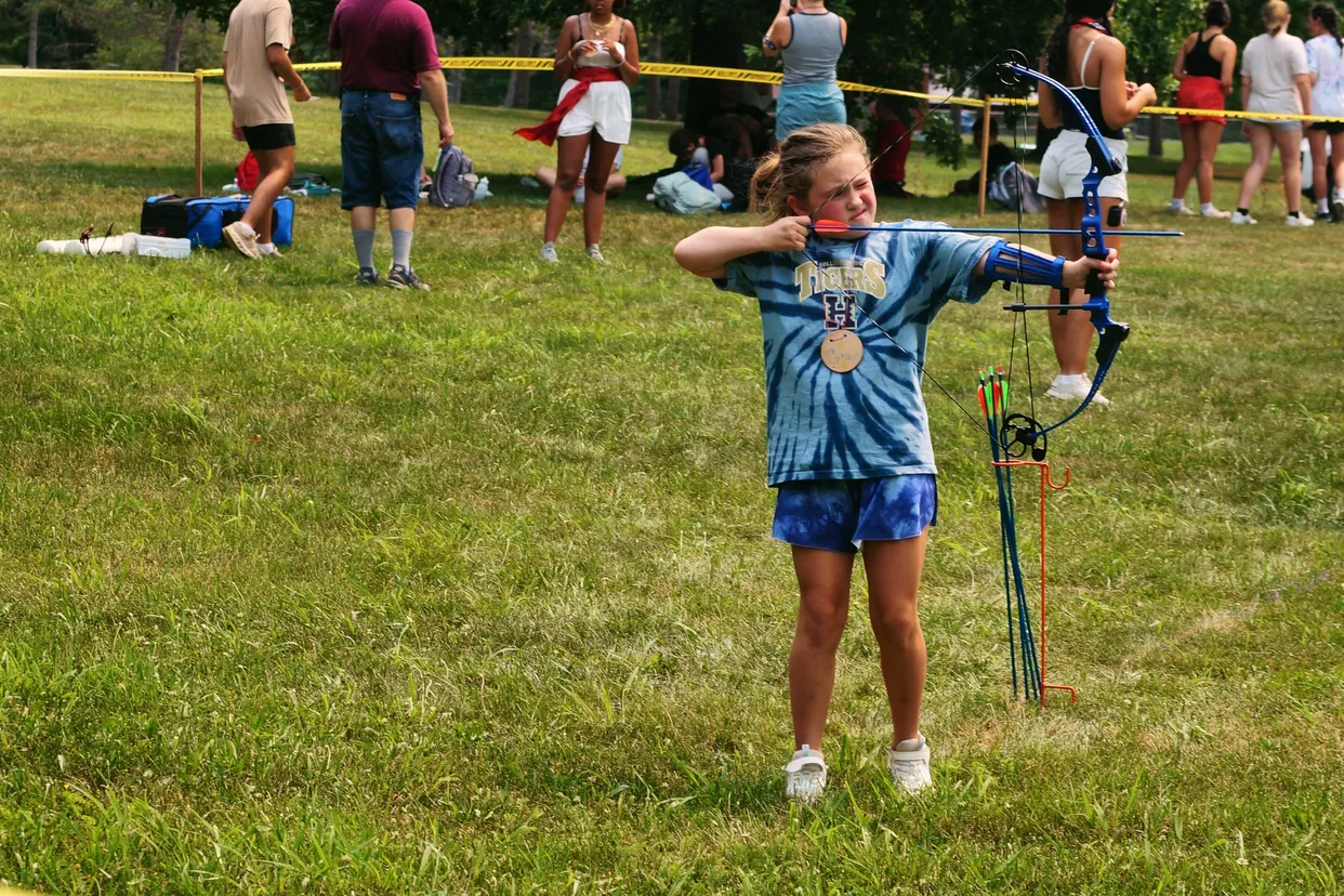 A girl in a tie-dye shirt aims a bow and arrow at a target on the archery field