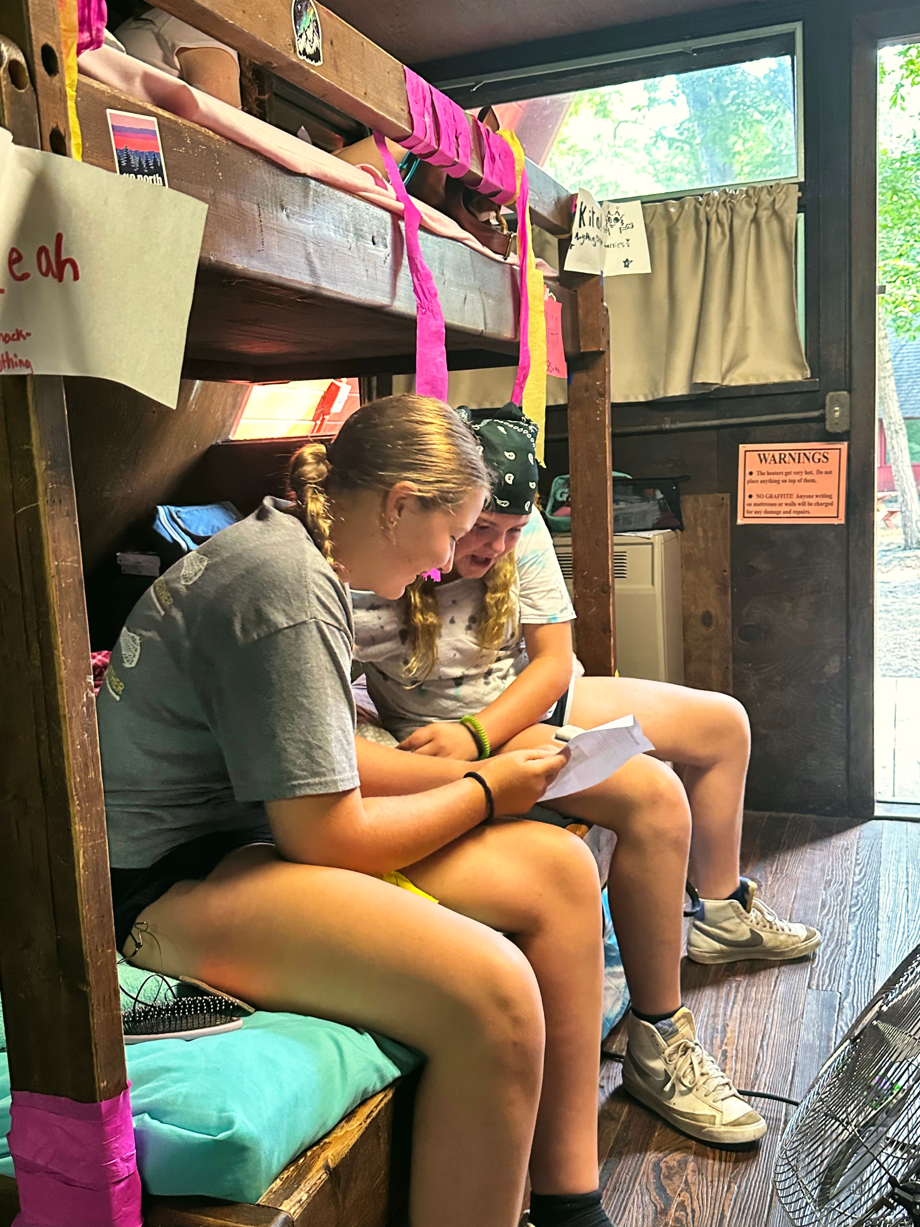 Two campers sit on a bunk bed in their cabin and read a note together.