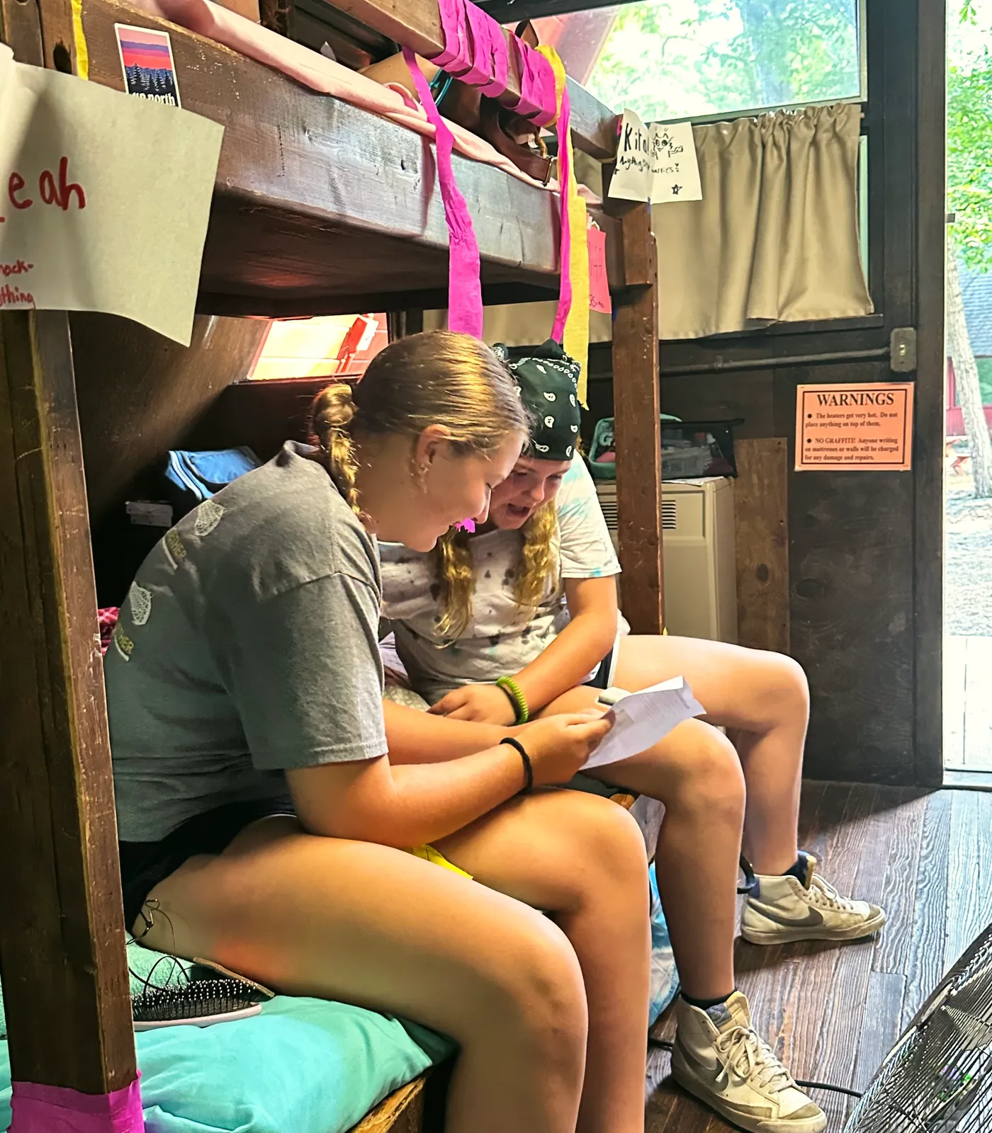 Two campers sit on a bunk bed in their cabin and read a note together.