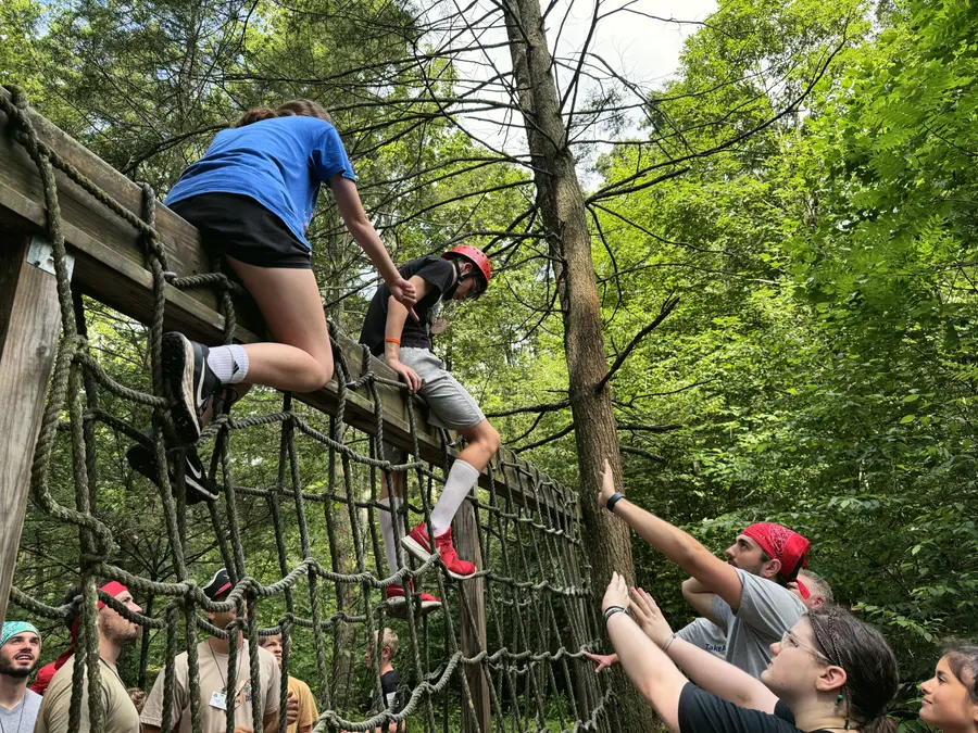 Campers help each other climb over a beam on the ropes course in the woods
