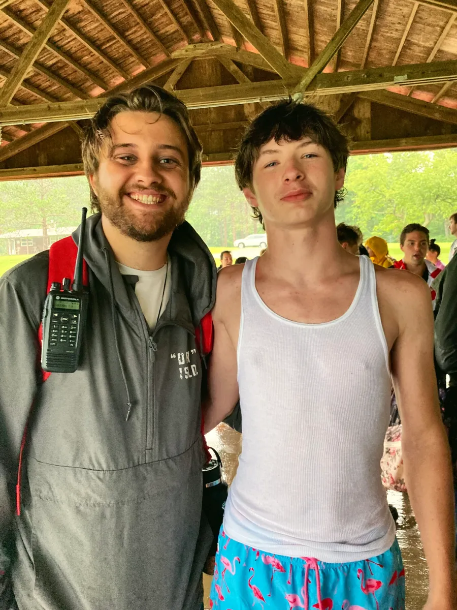 Two smiling young men under a wooden pavilion; one has a radio clipped to his jacket.