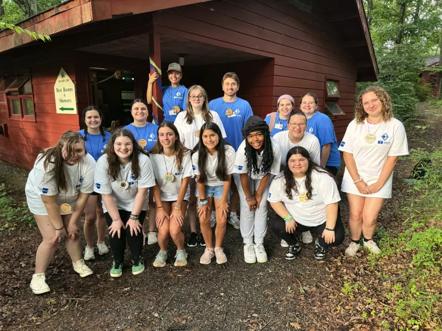 A group of teen campers smile together in front of a wooden cabin