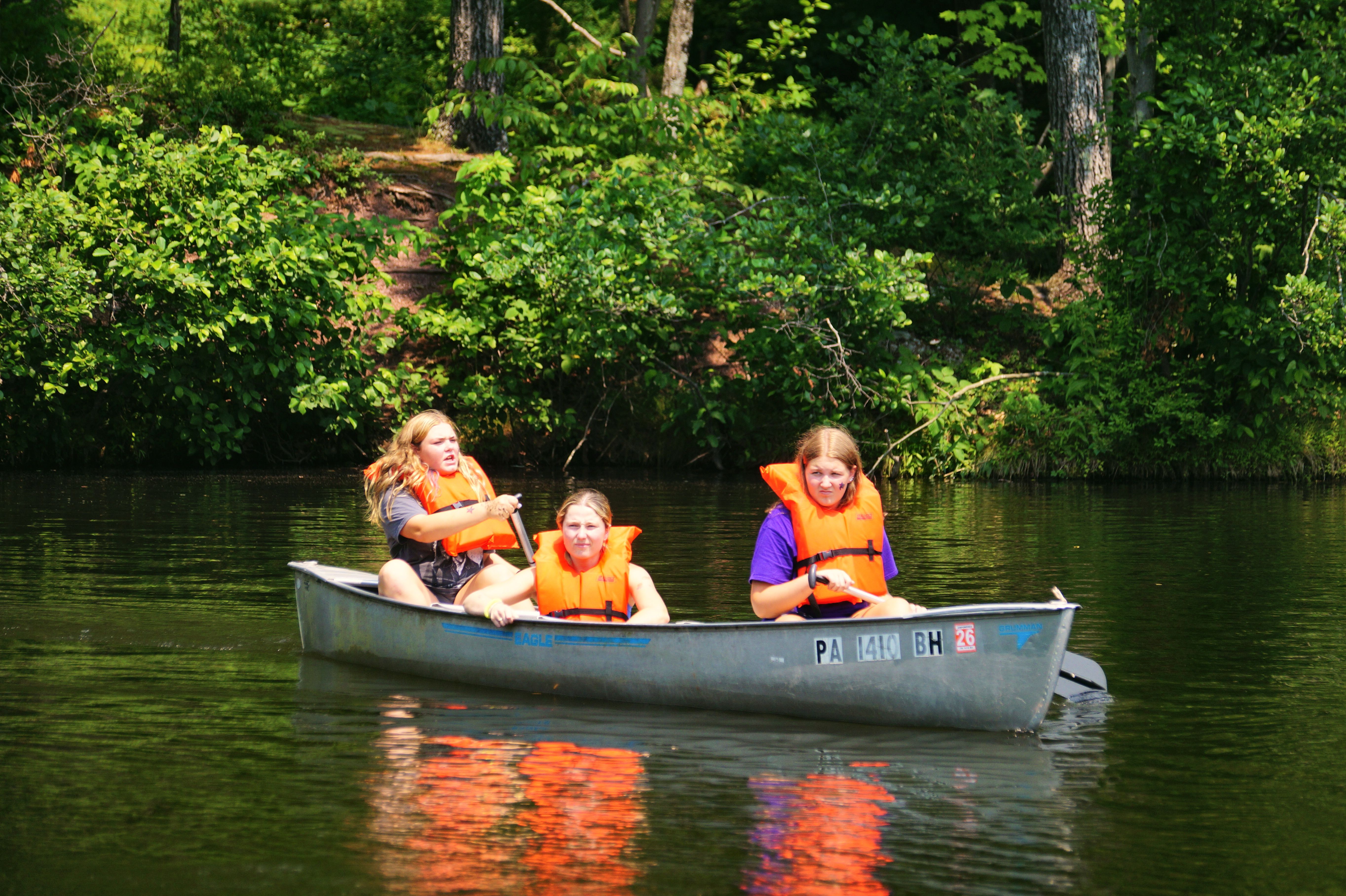 Three campers in orange life vests paddle a canoe across the camp lake.