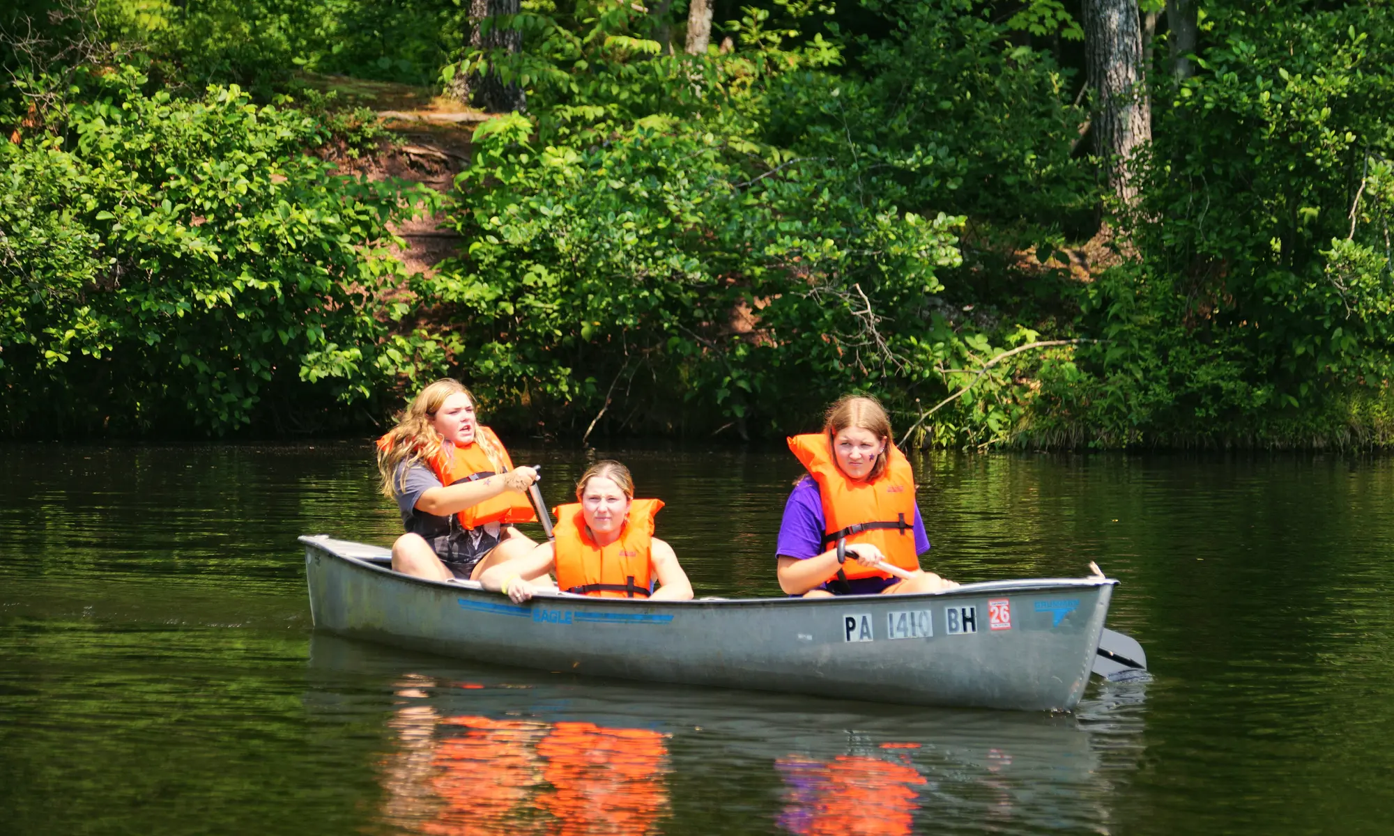 Three campers in orange life vests paddle a canoe across the camp lake.
