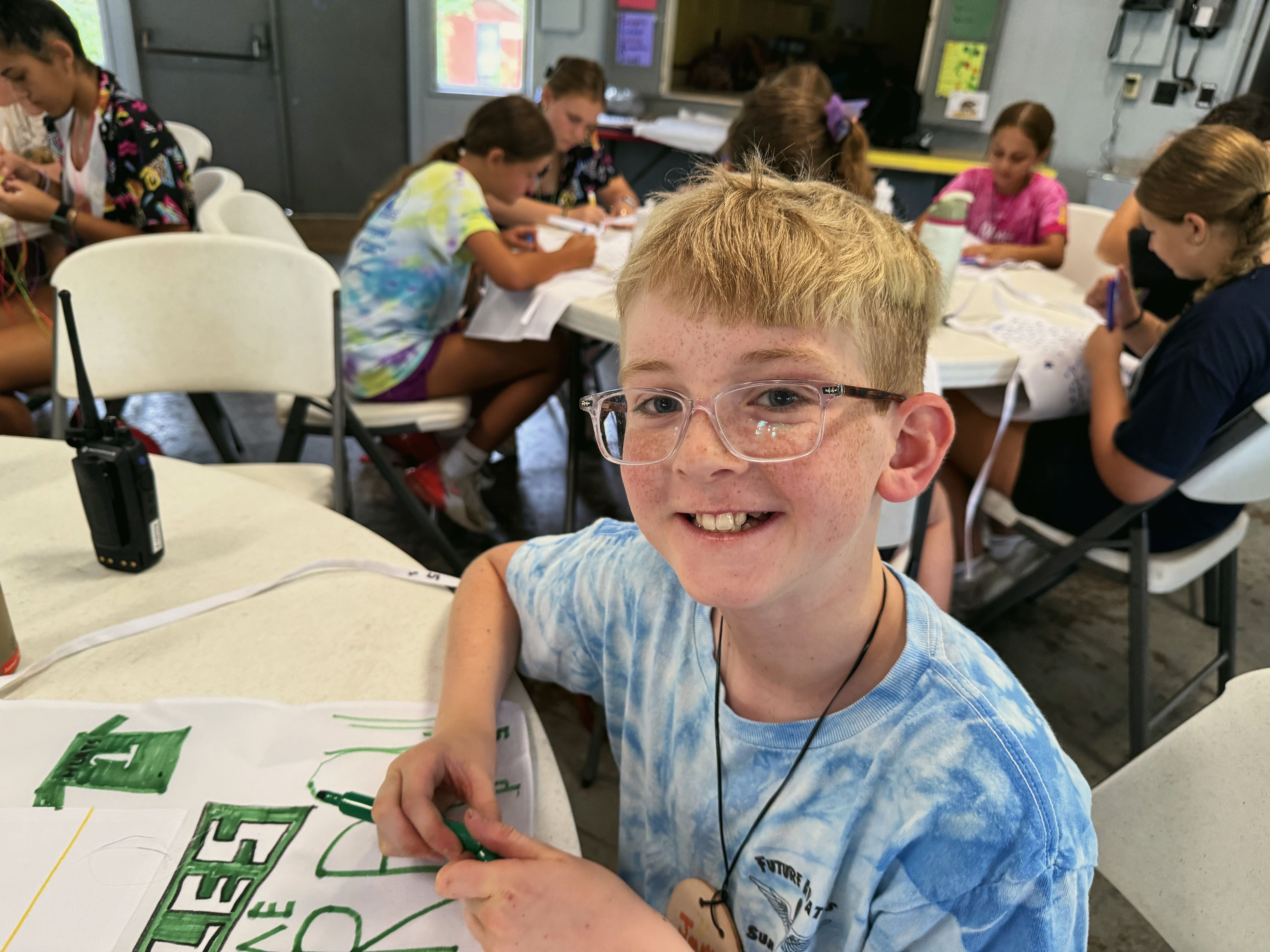 A young camper in glasses and a tie-dye shirt smiles at a drawing table with other children