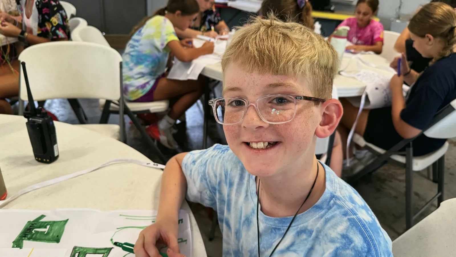 A young camper in glasses and a tie-dye shirt smiles at a drawing table with other children