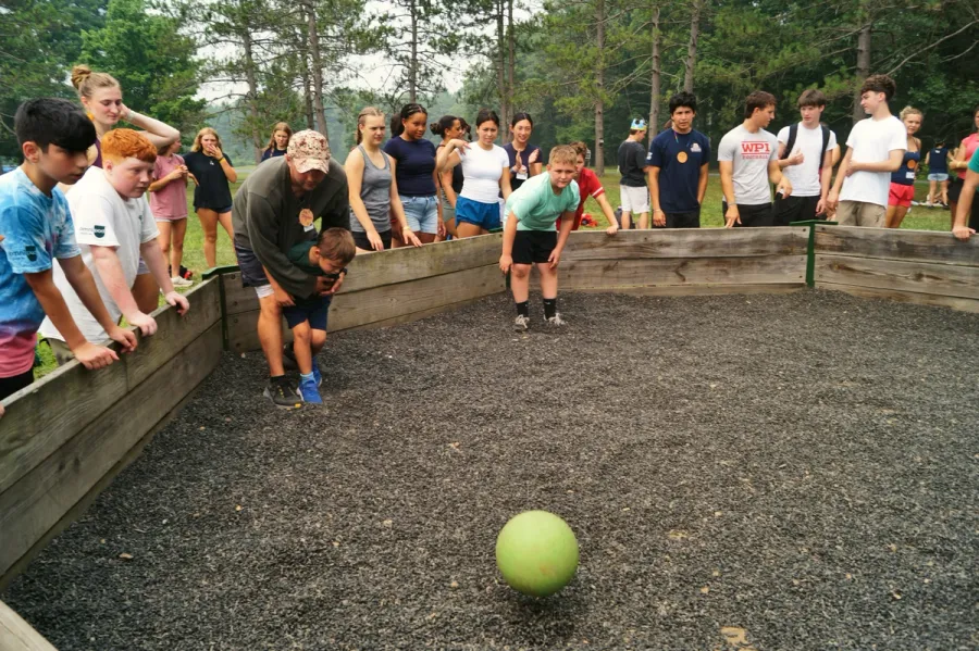 A man helps a young boy play a ball game in a gravel pit while teens and adults watch