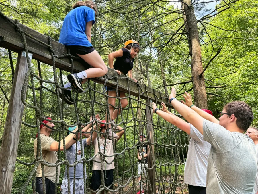 Teens help each other climb over a wooden beam on the ropes course in the woods