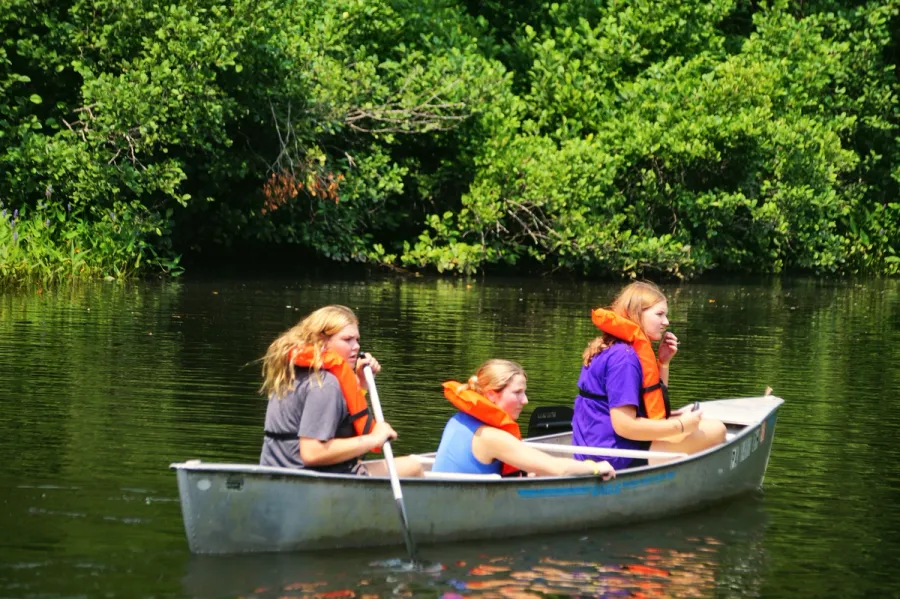 Three teen girls in orange life vests paddle a canoe on a calm river