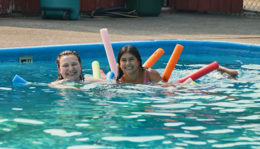 Two girls smile in the camp pool among colorful pool noodles.